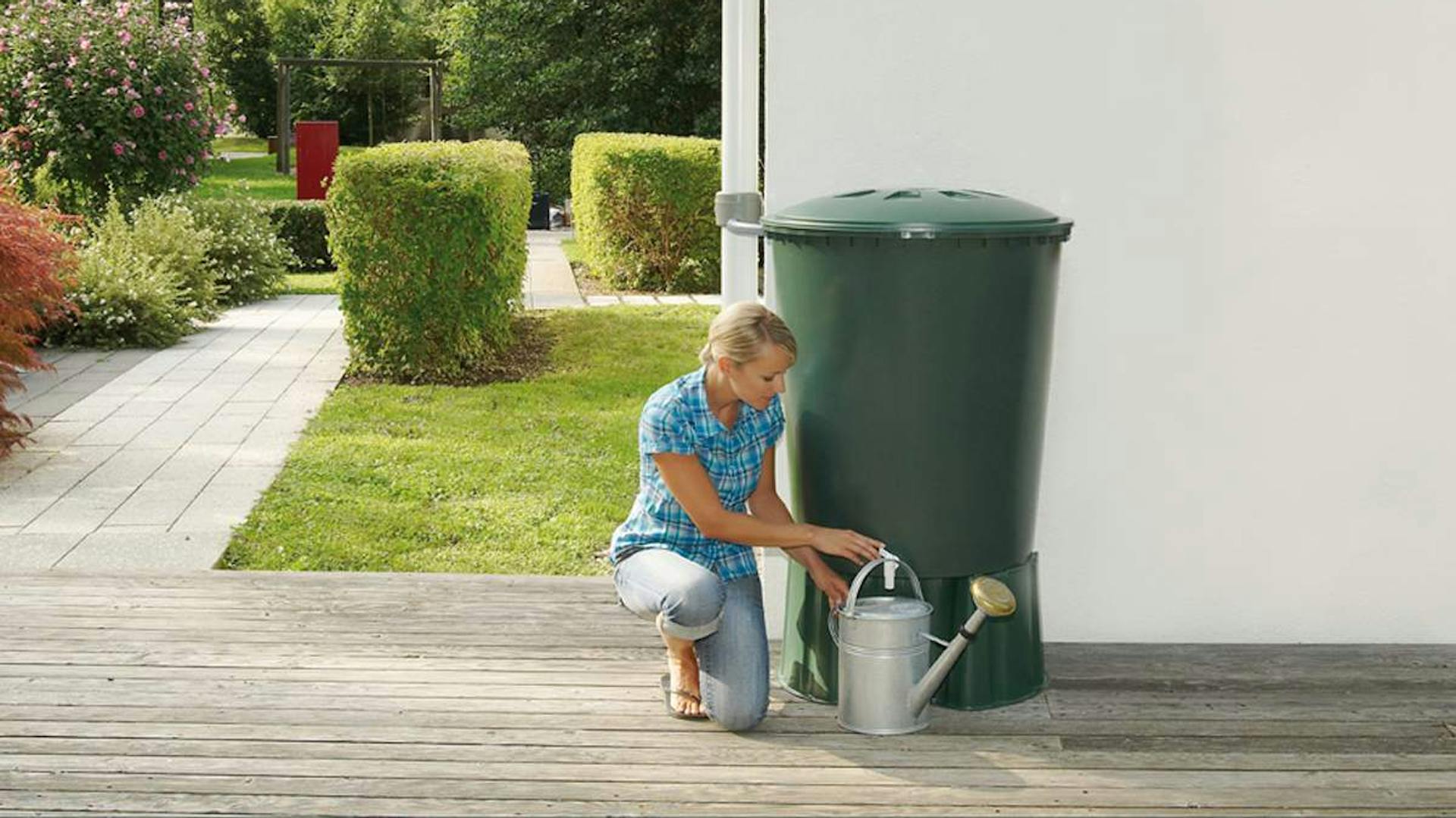 In un giardino accanto a una casa, una donna riempie un annaffiatoio prelevando l’acqua piovana da un serbatoio per l’acqua piovana
