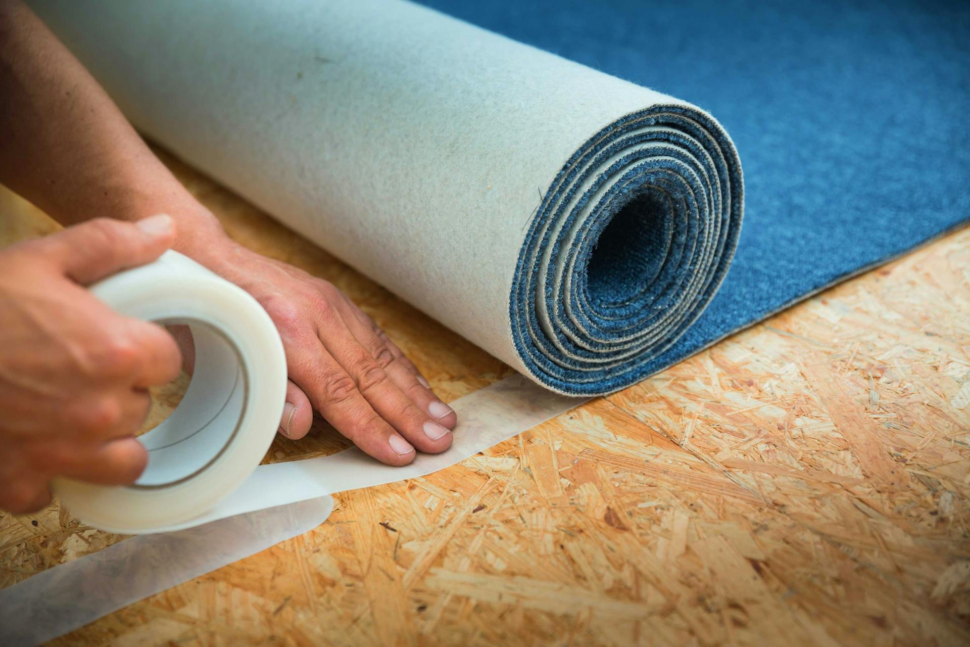 Pracovník fixuje modrý koberec k podlahe lepiacou páskou.