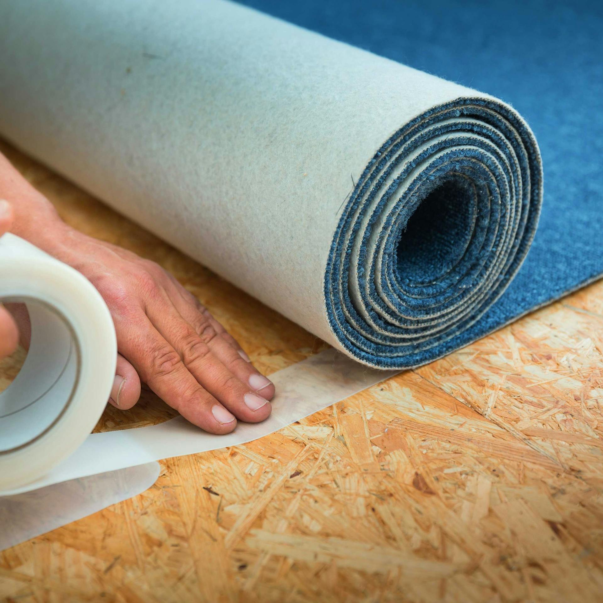 Heimwerker fixiert blauen Teppich mit Klebeband am Boden.