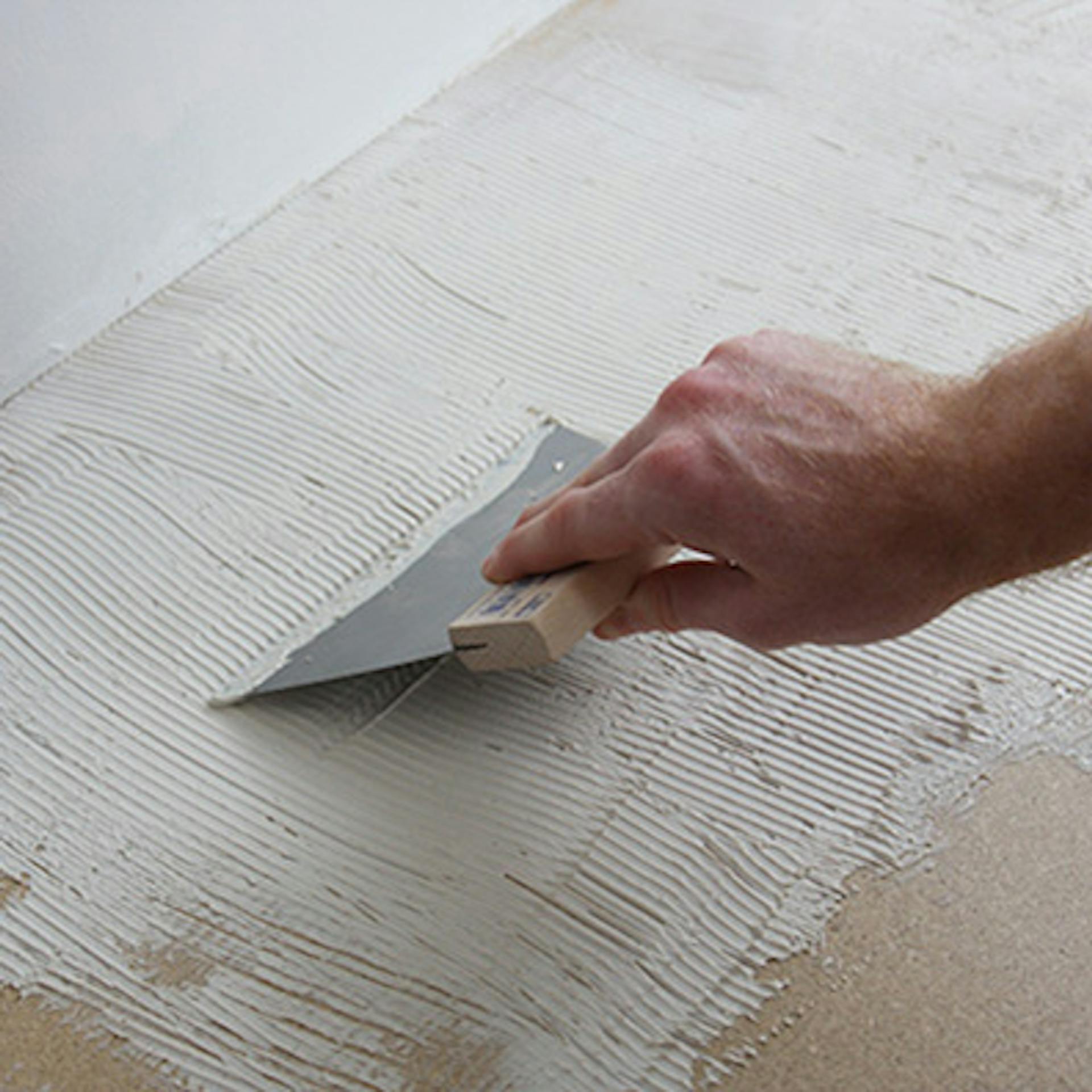 Heimwerker trägt Kleber mit Zahnspachtel auf Untergrund auf, um Teppich zu verlegen.