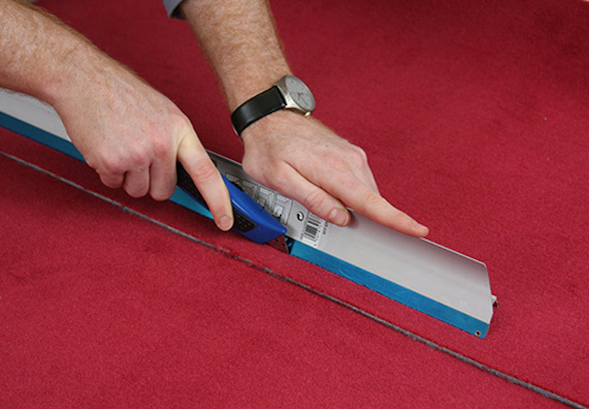 Pracovník ukladá druhý pás červeného koberca a orezáva koberec pomocou noža a pravítka.