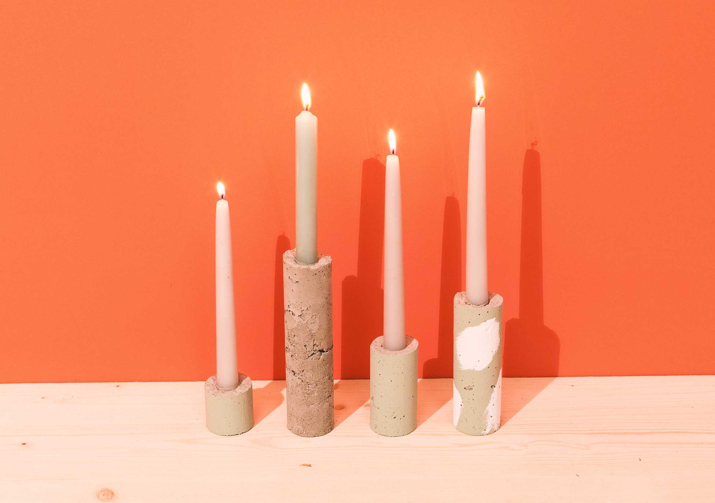 Lange, dünne Kerzen in unterschiedlich hohen Kerzenständern aus Beton vor orangener Wand