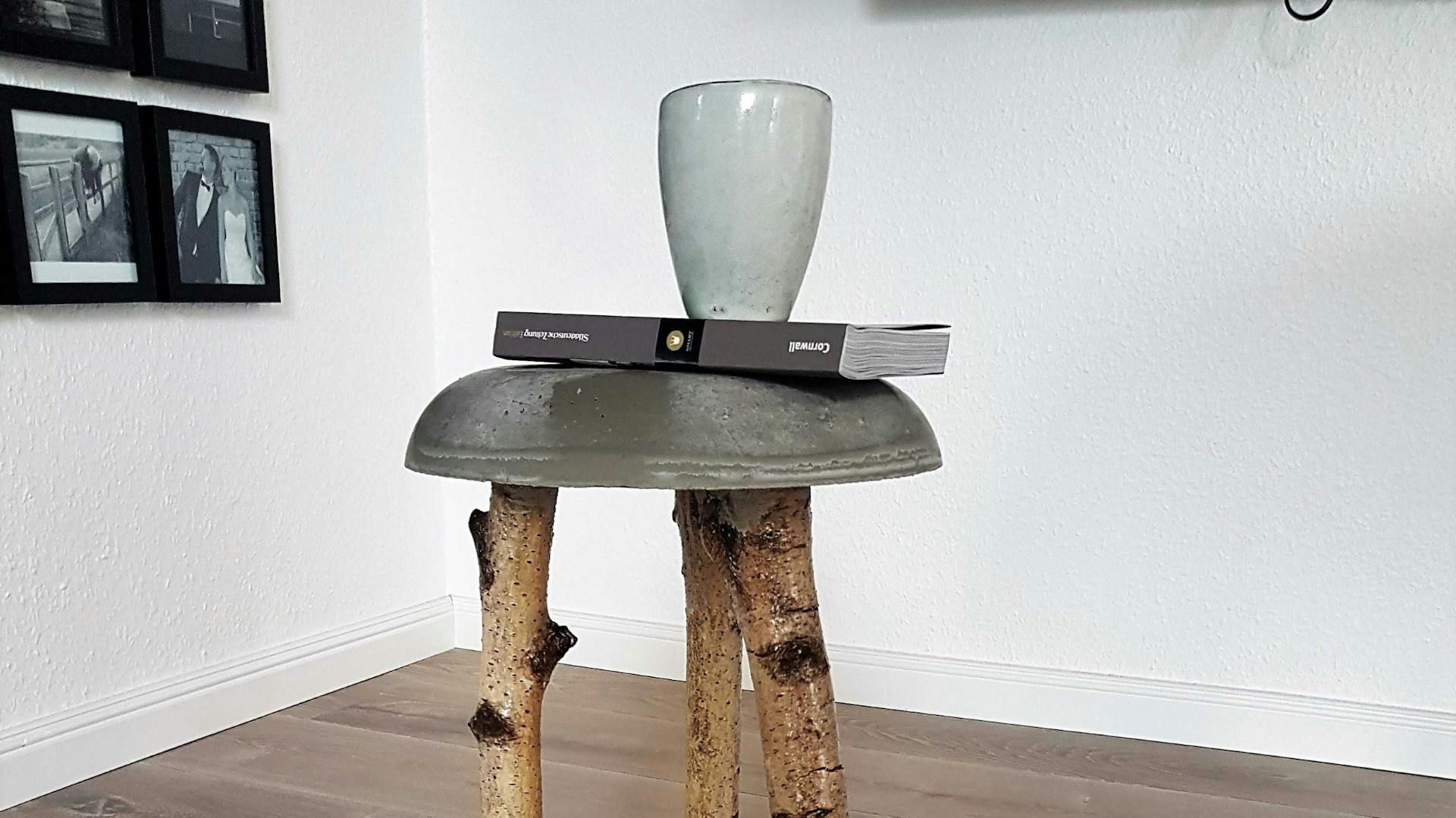 Hocker mit Sitzfläche aus Bastelbeton mit Buch und Becher und Füßen aus Ästen auf Holzboden mit Lammfell