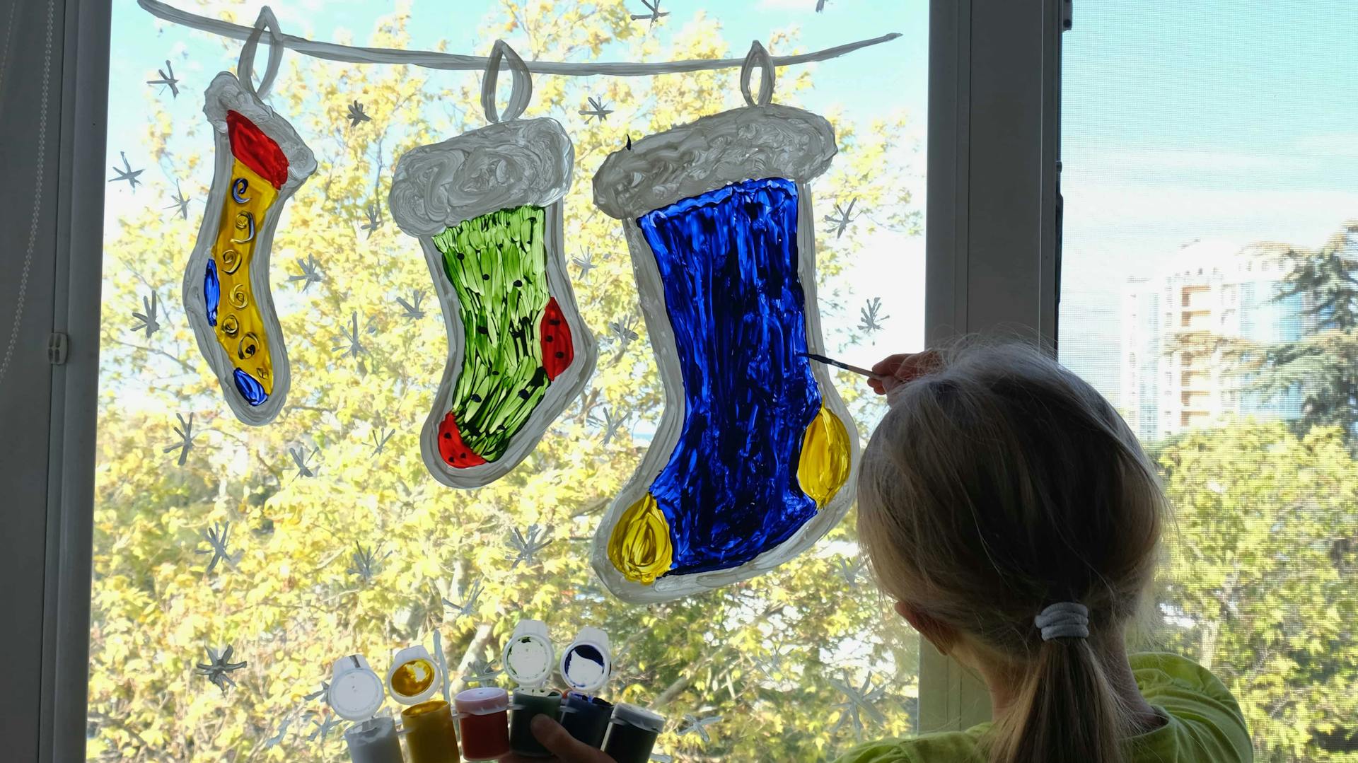 Ein kleines Mädchen malt mit Fensterfarbe Weihnachtssocken an eine Fensterscheibe.