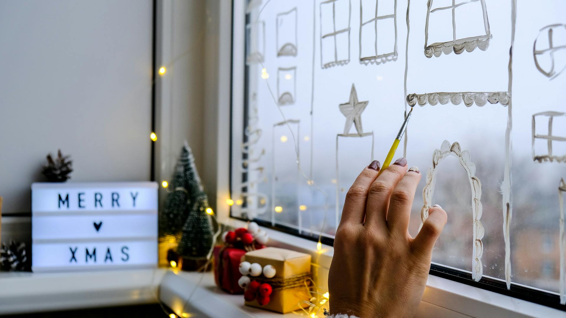 Jemand malt eine Winterstadt mit Fensterfarbe auf ein Fenster.