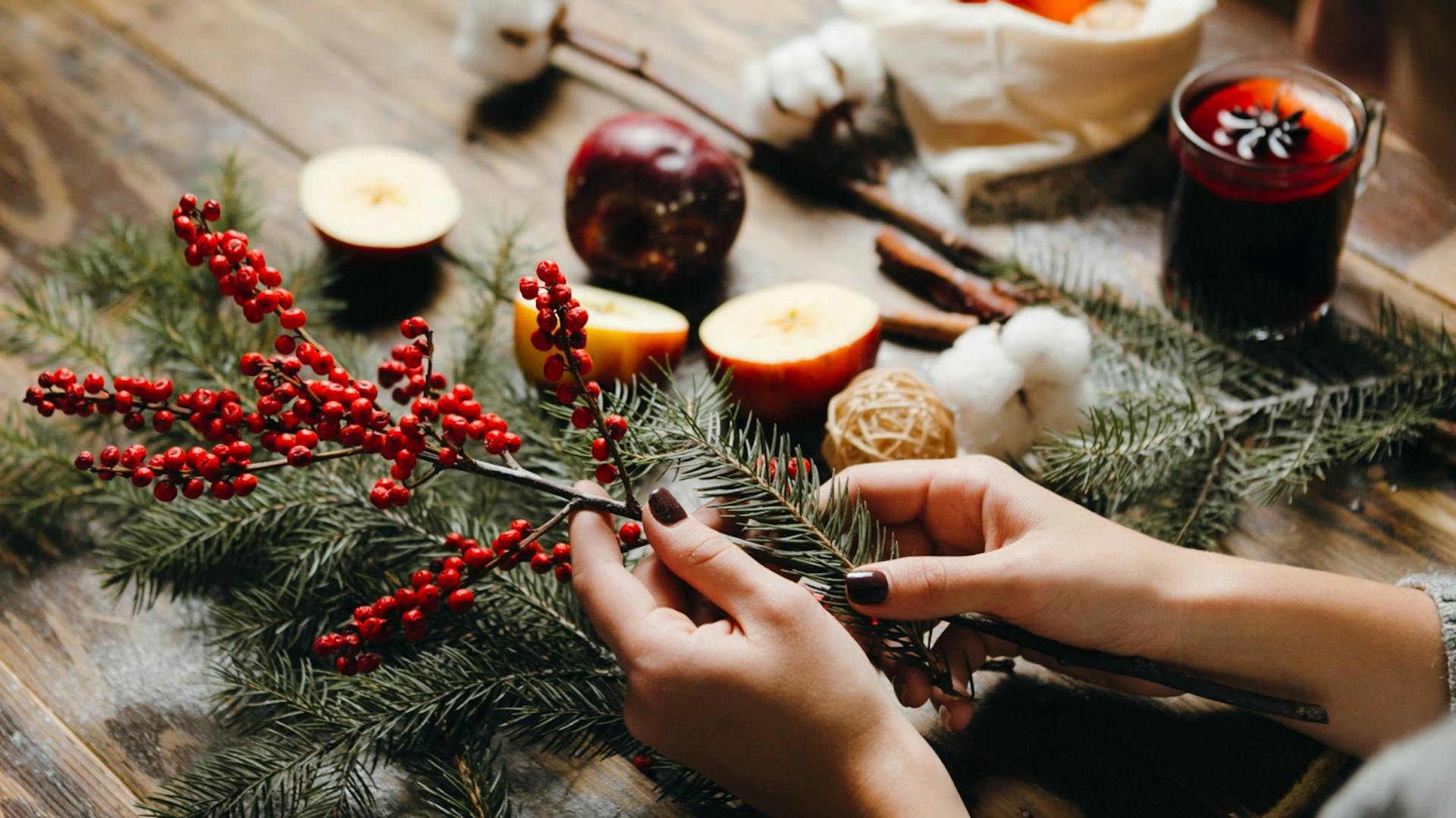 Una persona realizza una corona dell’Avvento con ramoscelli d’abete e altre piante. Sul tavolo poggiano mele tagliate a metà, accanto c’è un bicchiere di vin brulé.