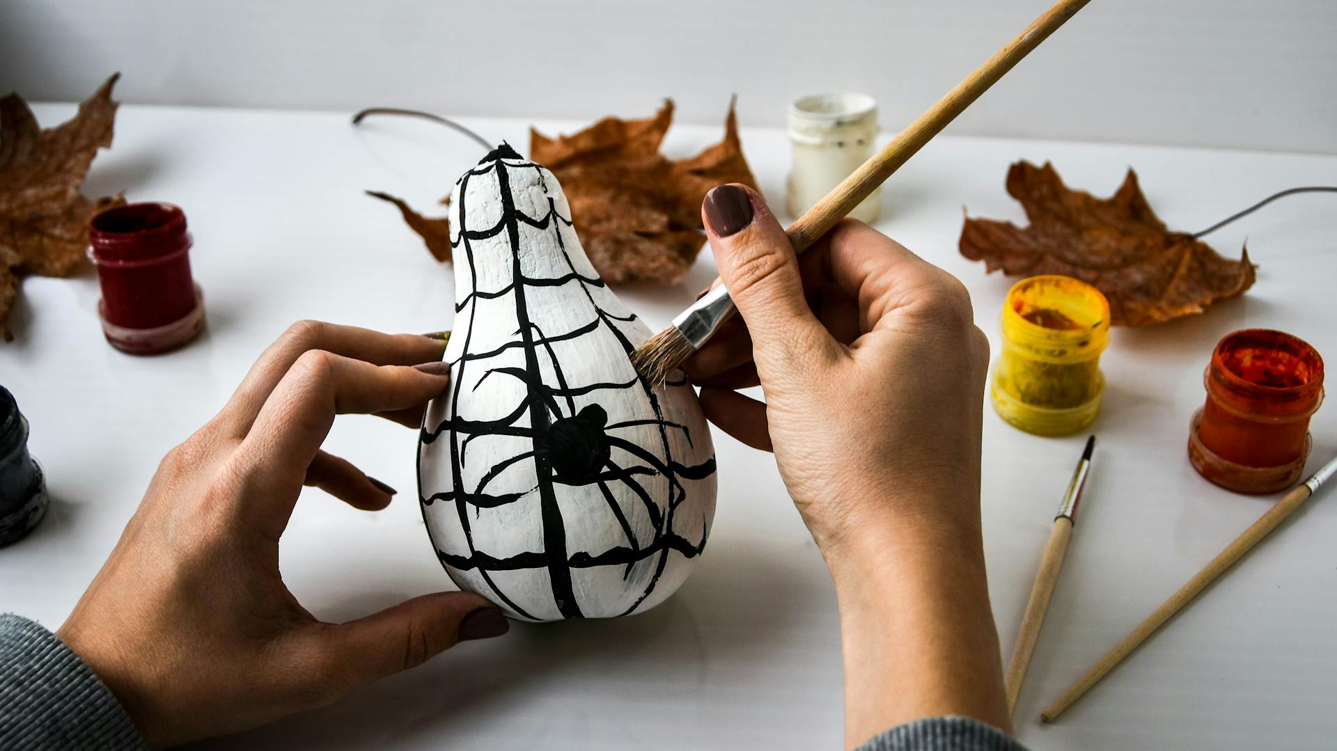 Jemand bemalt einen Kürbis mit weißer Farbe und einem dunklen Spinnennetz.