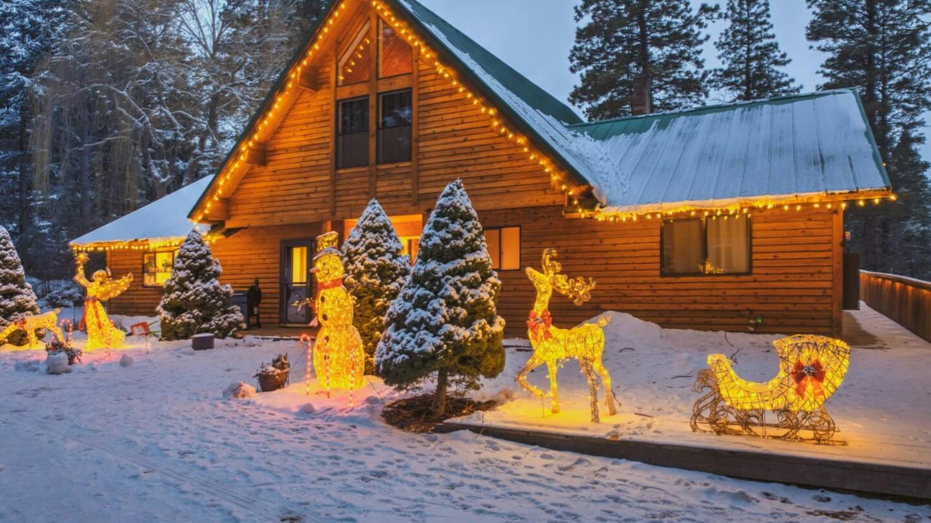 Grande casa in legno. Il bordo del tetto è decorato con ghirlande luminose. Nel giardino innevato ci sono figure luminose.