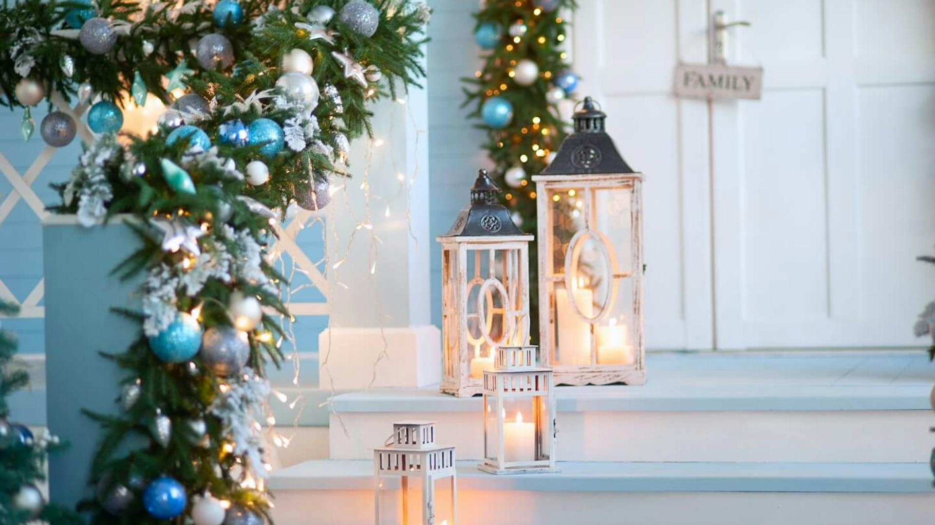 Scala per esterni bianca per la porta d’ingresso. Ringhiera decorata con ramoscelli d’abete e palline di Natale blu, accanto ci sono tre lanterne con candela.