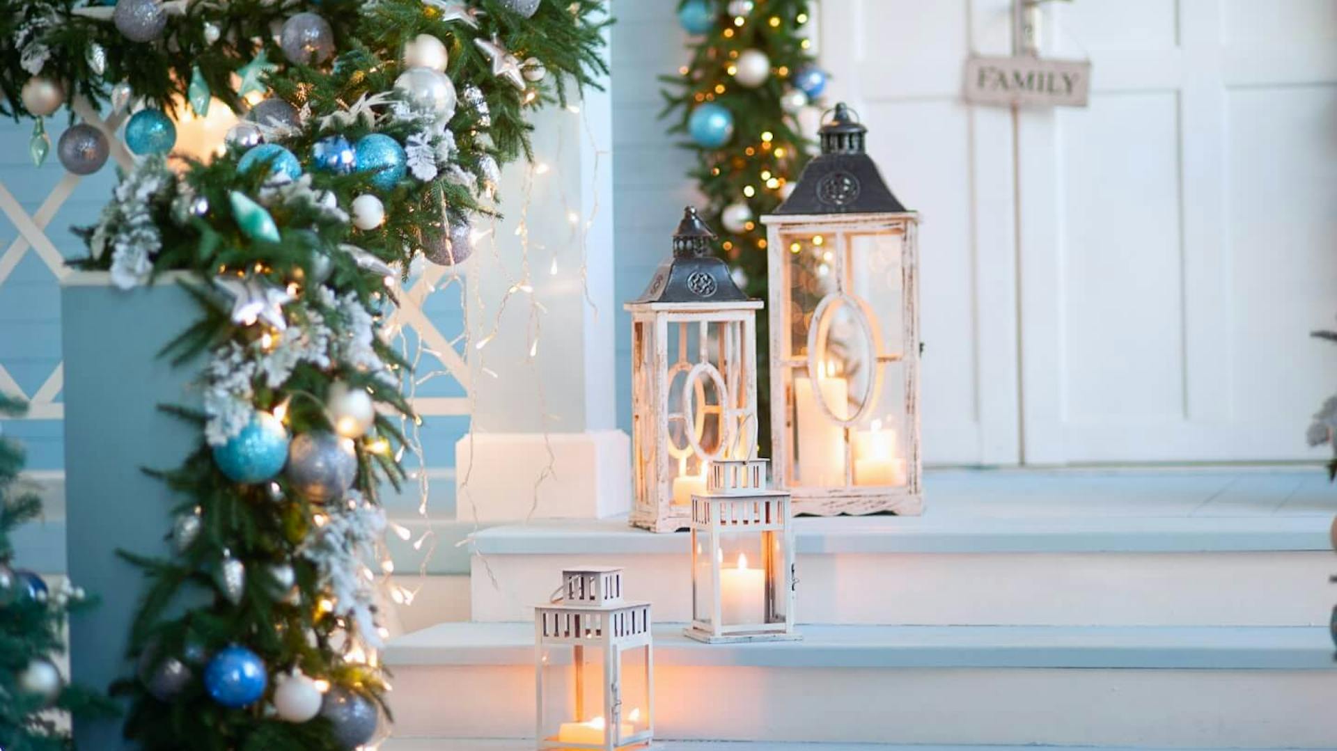 Weiße Außentreppe zur Eingangstür. Mit Tannenzweigen und blauen Weihnachtskugeln verziertes Geländer, daneben stehen drei Laternen mit Kerze.