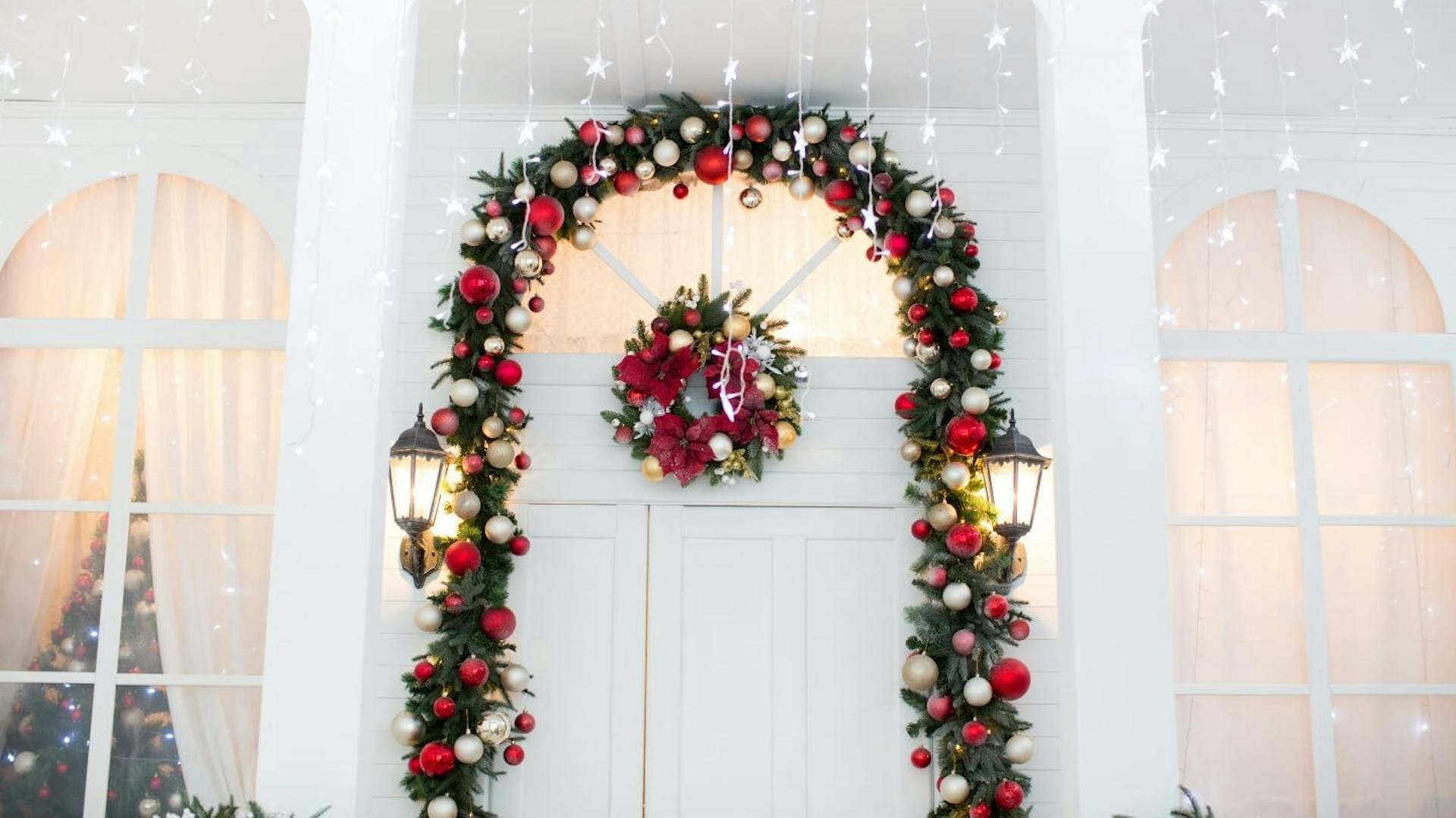 Weiße Eingangstür geschmückt mit einem Bogen aus Tannenzweigen und weißen und silbernen Weihnachtskugeln. Darüber hängen Lichterketten in Sternenform