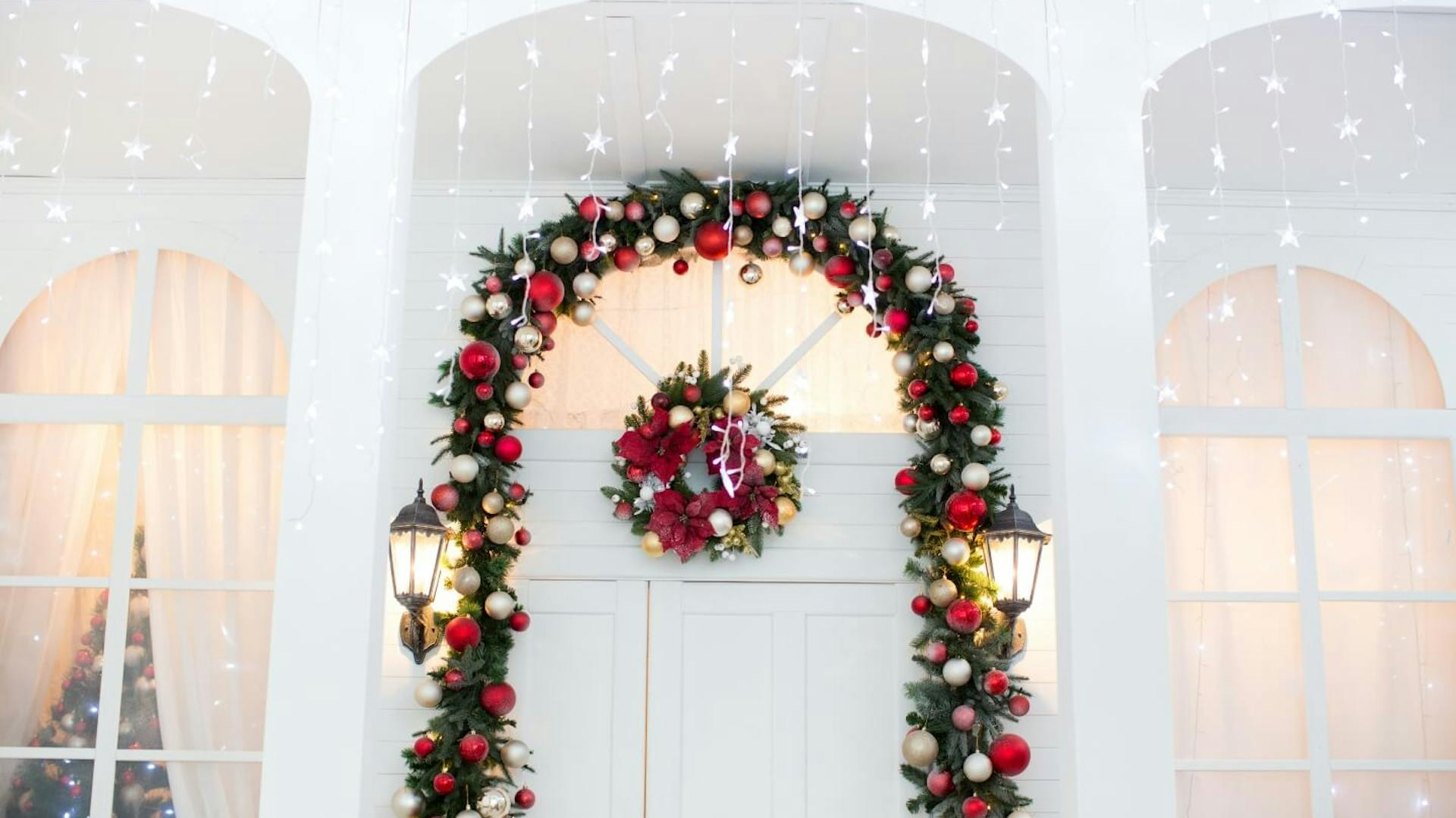 Porta d’ingresso bianca decorata con un arco di ramoscelli d’abete e palline di Natale color bianco e argento. Sopra di esso pendono ghirlande luminose a forma di stella
