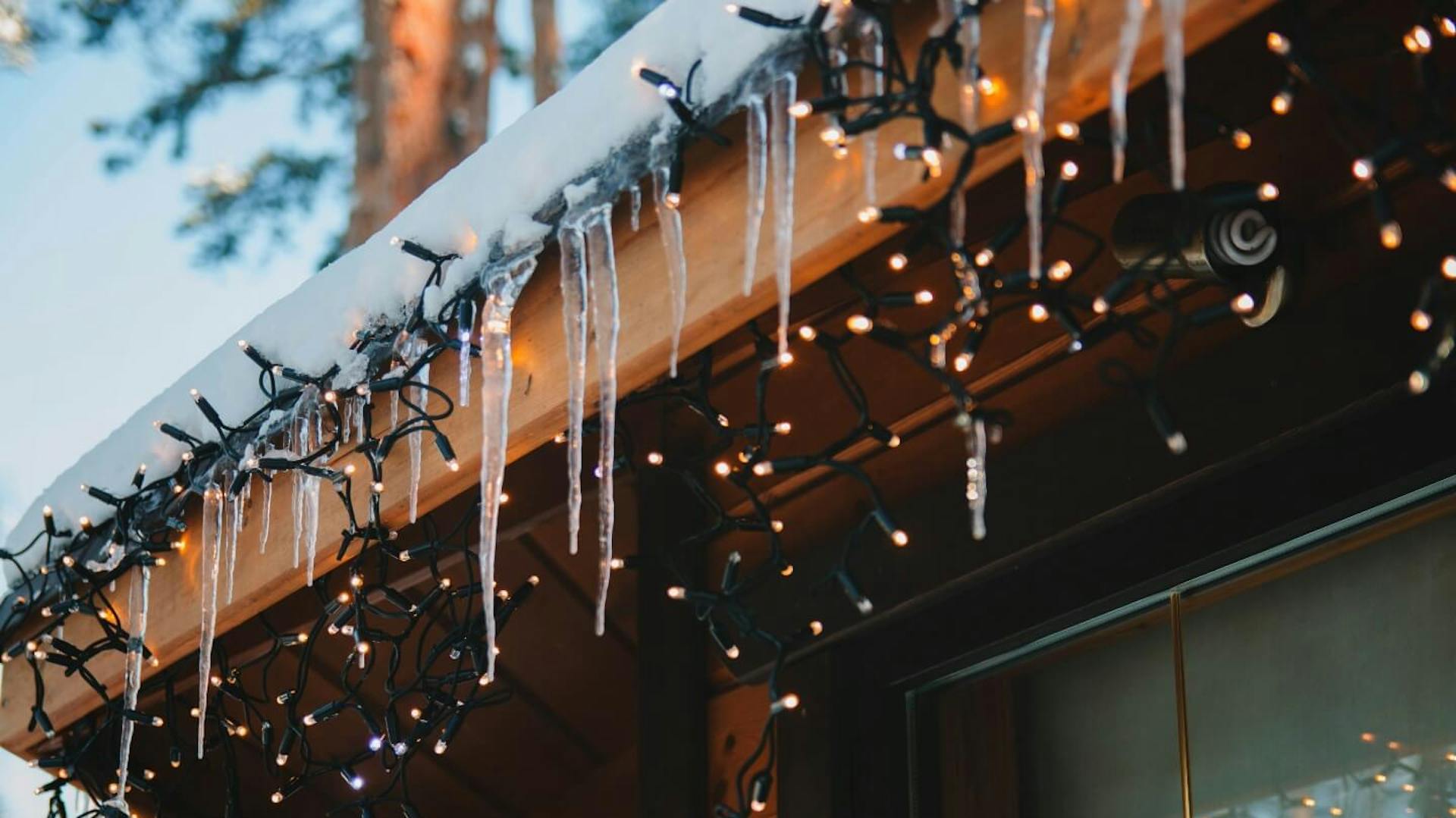 Außenkante eines schneebedeckten Dachs mit Eiszapfen. Daran ist eine Lichterkette angebracht.
