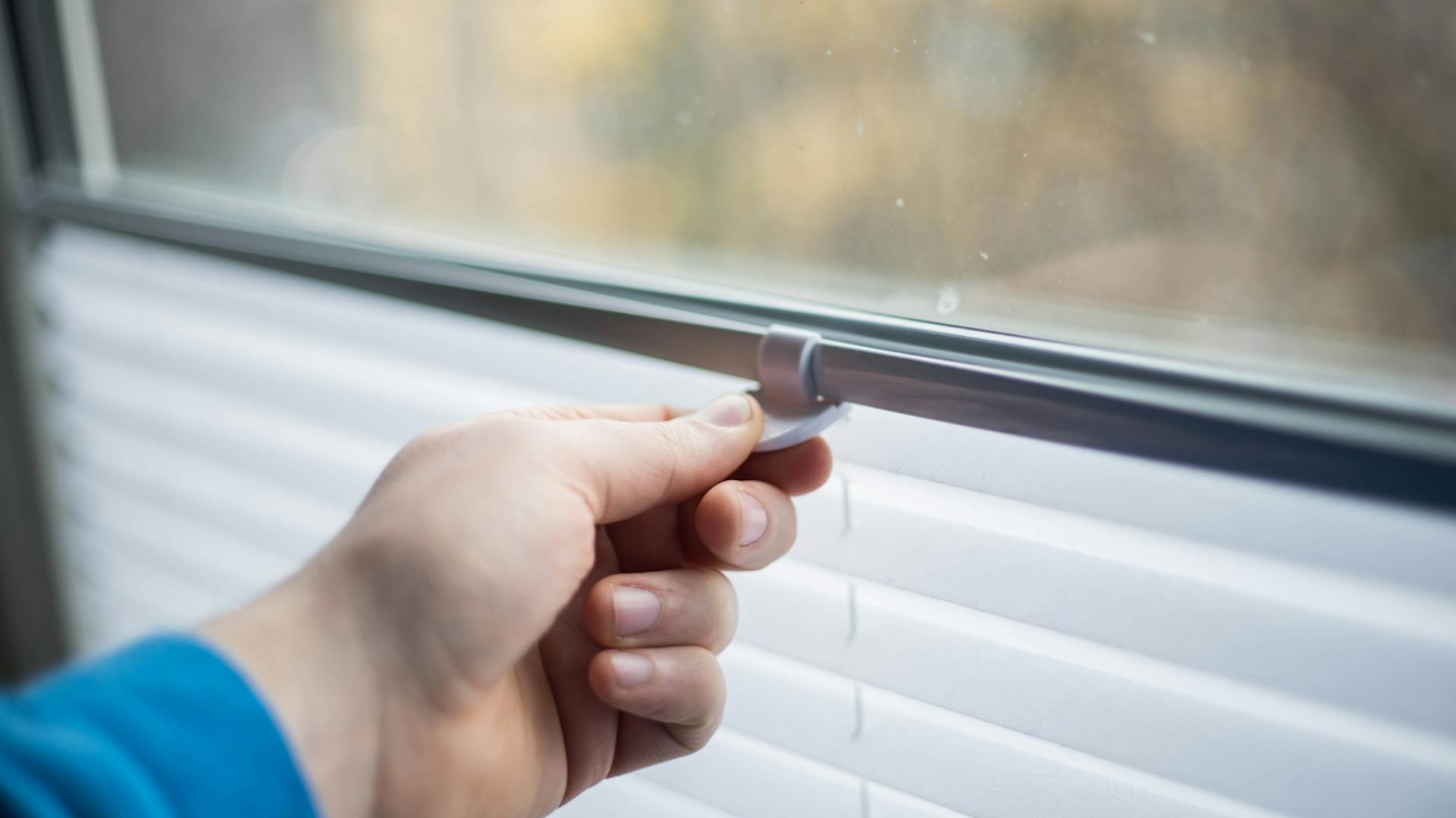 Es ist ein Fenster zu sehen. Eine Person hält die Halterung eines Plissees mit der Hand fest.