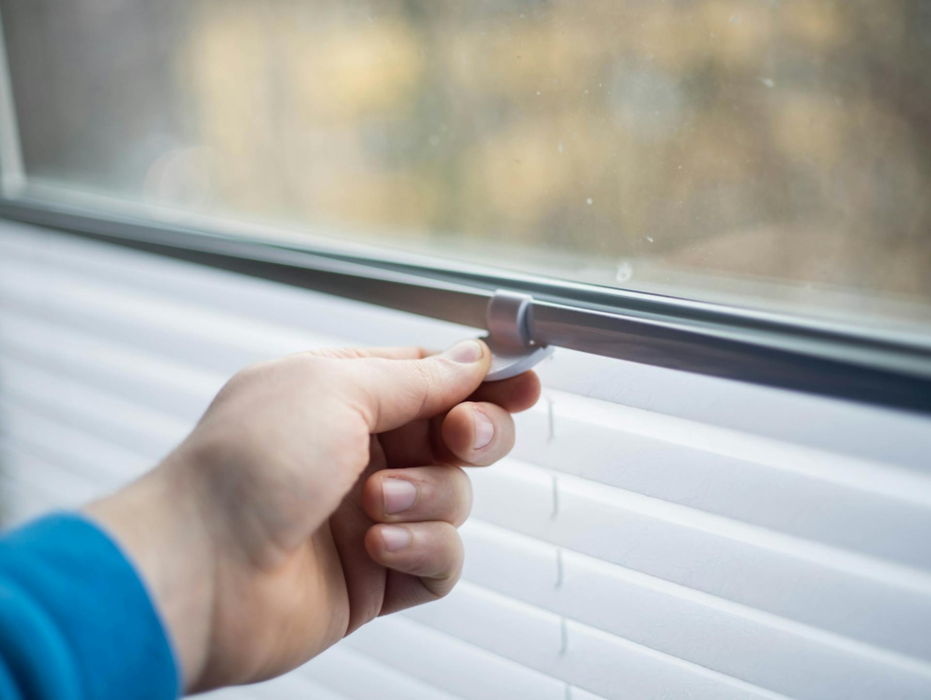 La figura mostra una finestra. Una persona tiene saldamente in mano il supporto di una tenda plissettata.