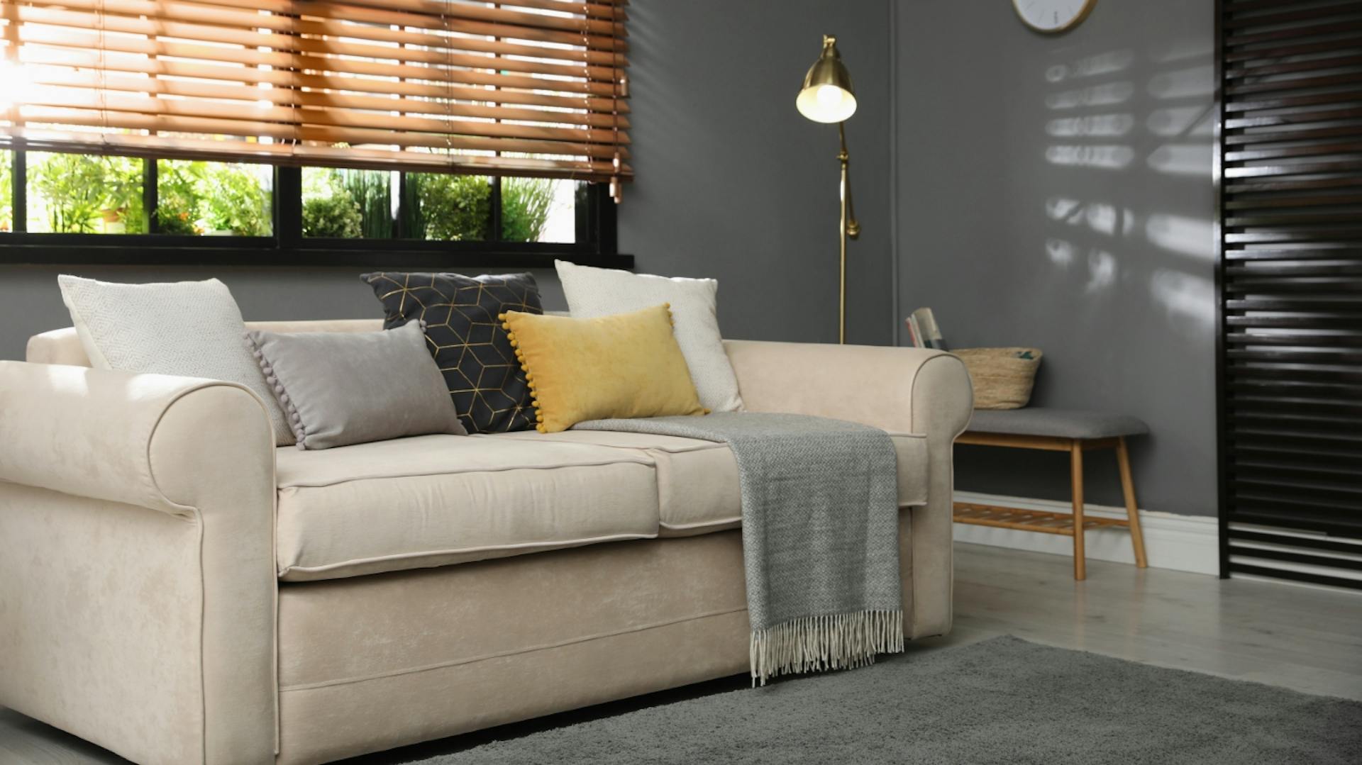 Es ist eine beige Couch zu sehen, welche vor einer grauen Wand steht. Auf der Couch liegen fünf Kissen und eine Decke. Neben der Couch stehen eine goldene Stehlampe und eine kleine Bank mit Holzfüßen. An der Wand hängt eine Uhr. Hinter der Couch ist ein Fenster mit dunklen Sprossen zu sehen, davor hängt eine heruntergelassene, hölzerne Jalousie. Vor der Couch liegt ein grauer Teppich.
