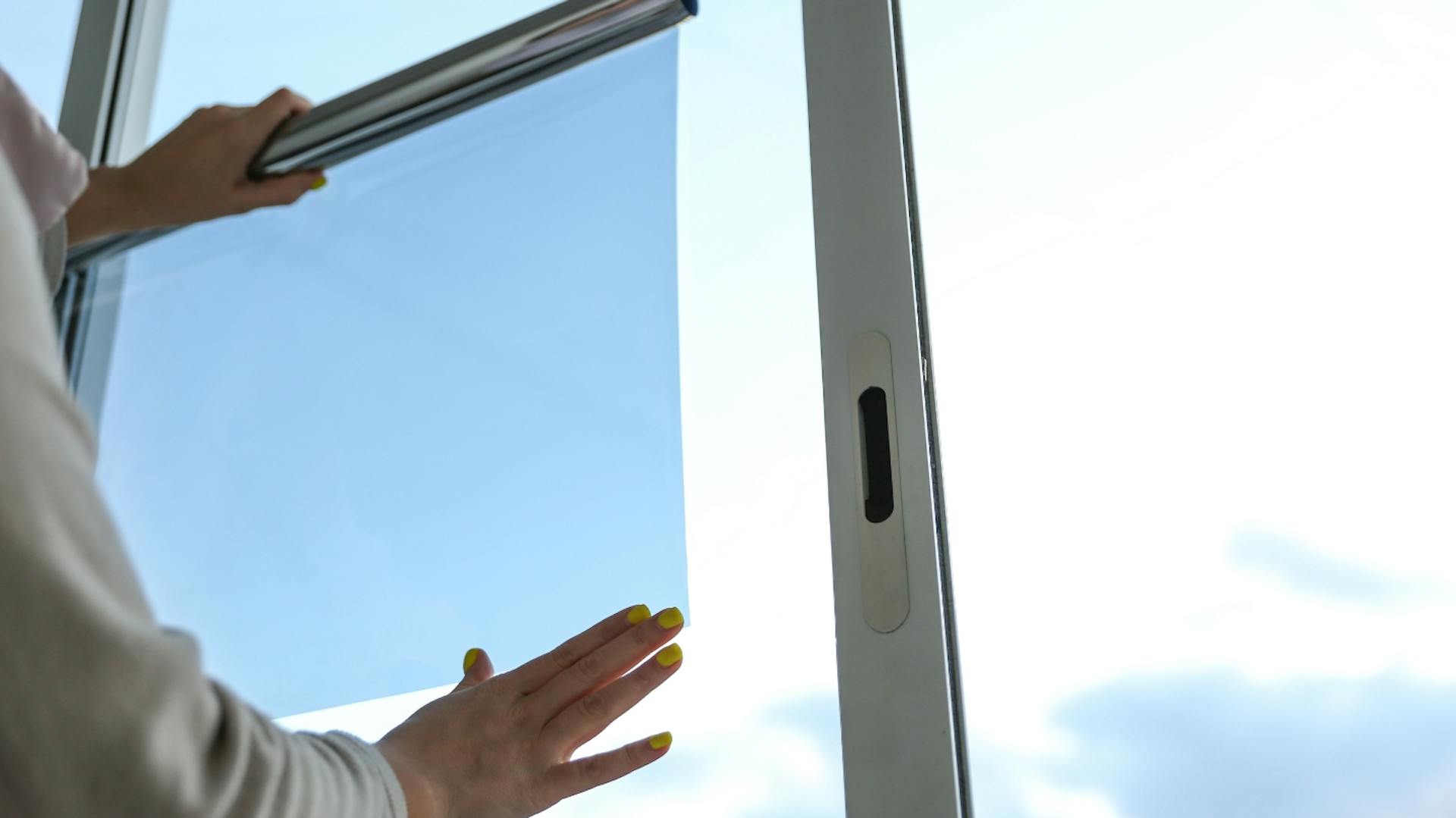 Es ist ein Fenster zu sehen. Davor steht ein Person, welche eine Sonnenschutzfolie an dem Fenster anbringt. Es sind nur die Hände und Arme zu sehen.