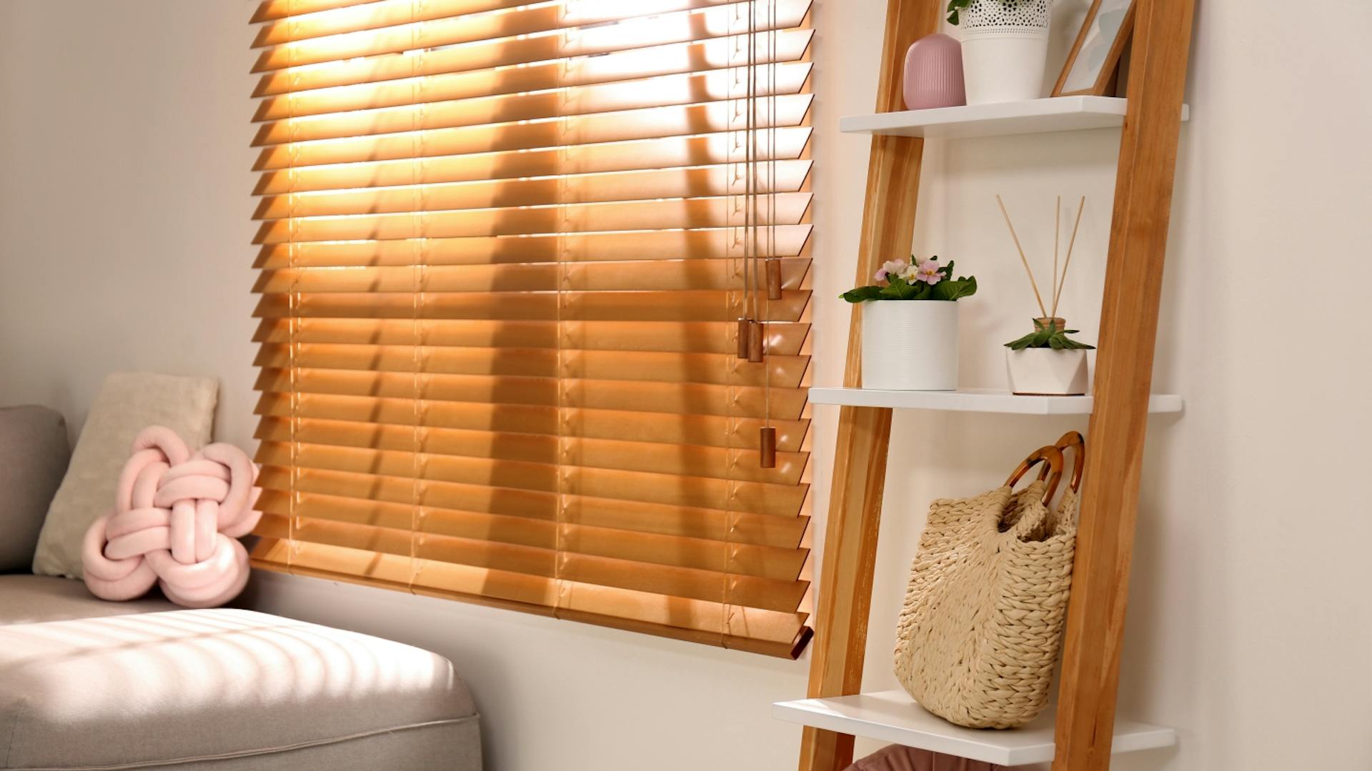 Es ist ein Teil von einem grauen Sofa zu sehen. An der Wand steht ein Regal mit Holzstreben und weißen Regalböden. Auf den einzelnen Böden stehen kleine Pflanzen sowie eine Tasche. Ein Fenster wird durch eine heruntergelassene Jalousie aus Holz verdeckt.