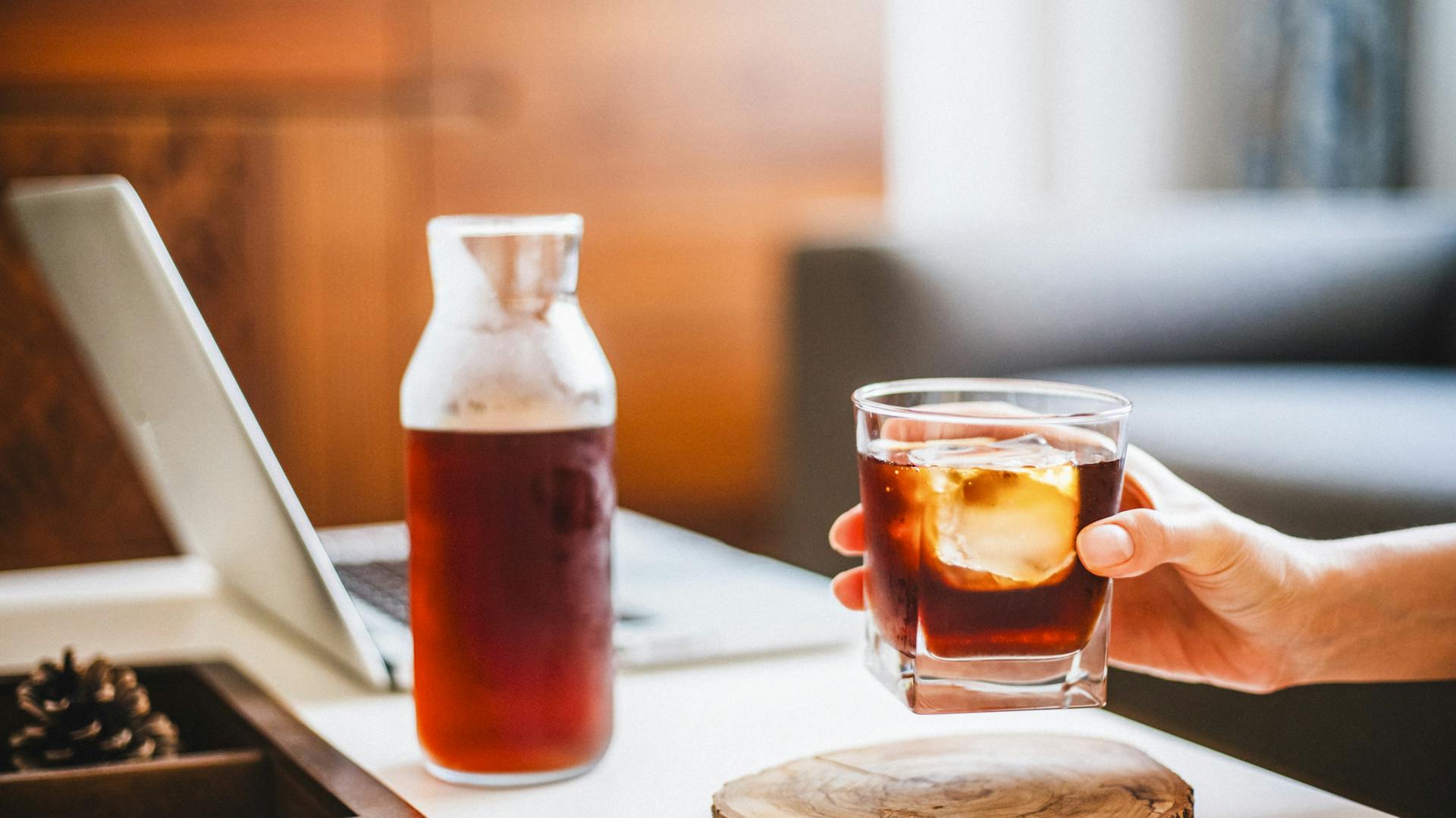 Person stellt ein Glas Cold-Brew-Coffee neben einem Notebook ab