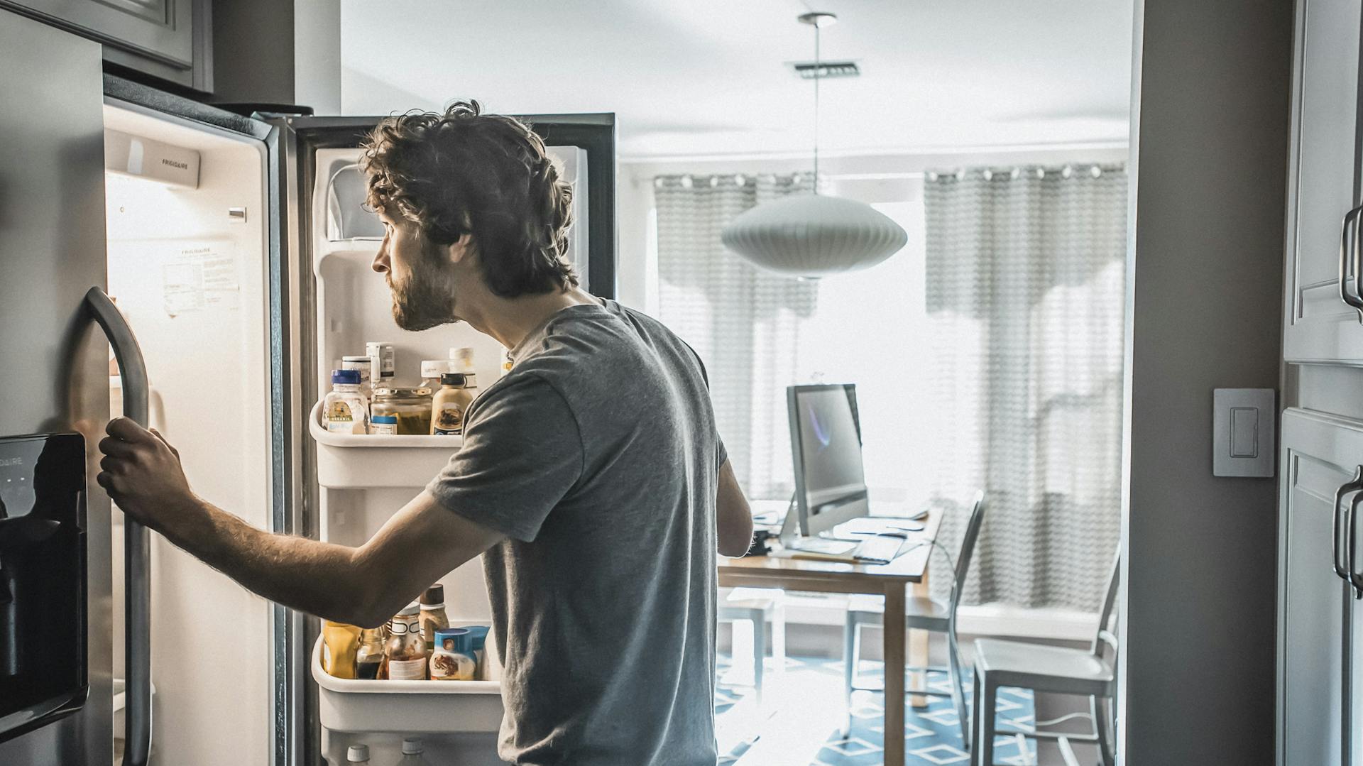 Mann schaut in den Kühlschrank, im Hintergrund sein Homeoffice