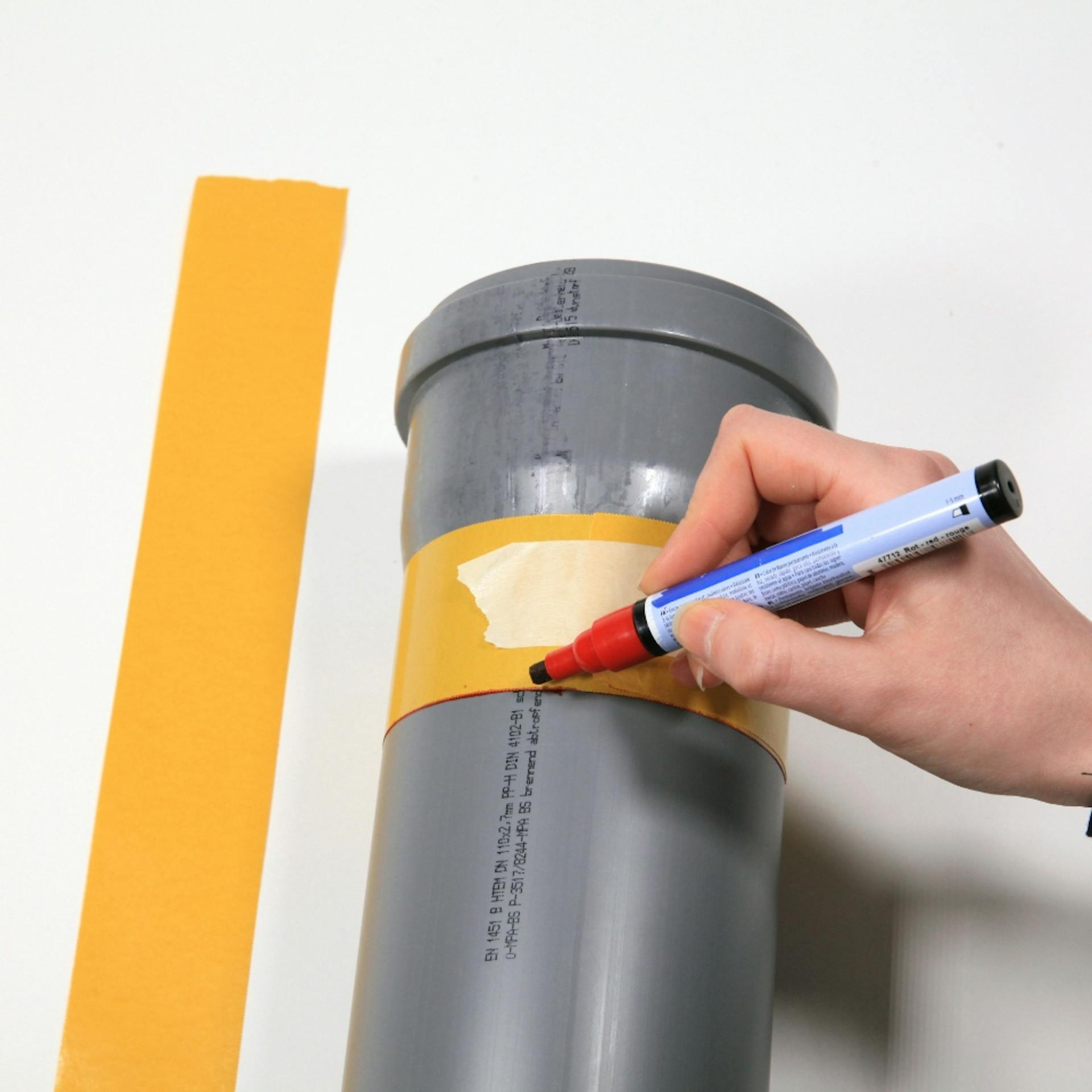 Kreppband klebt um Abflussrohr. Person zeichnet Linie entlang des Bands mit rotem Marker.