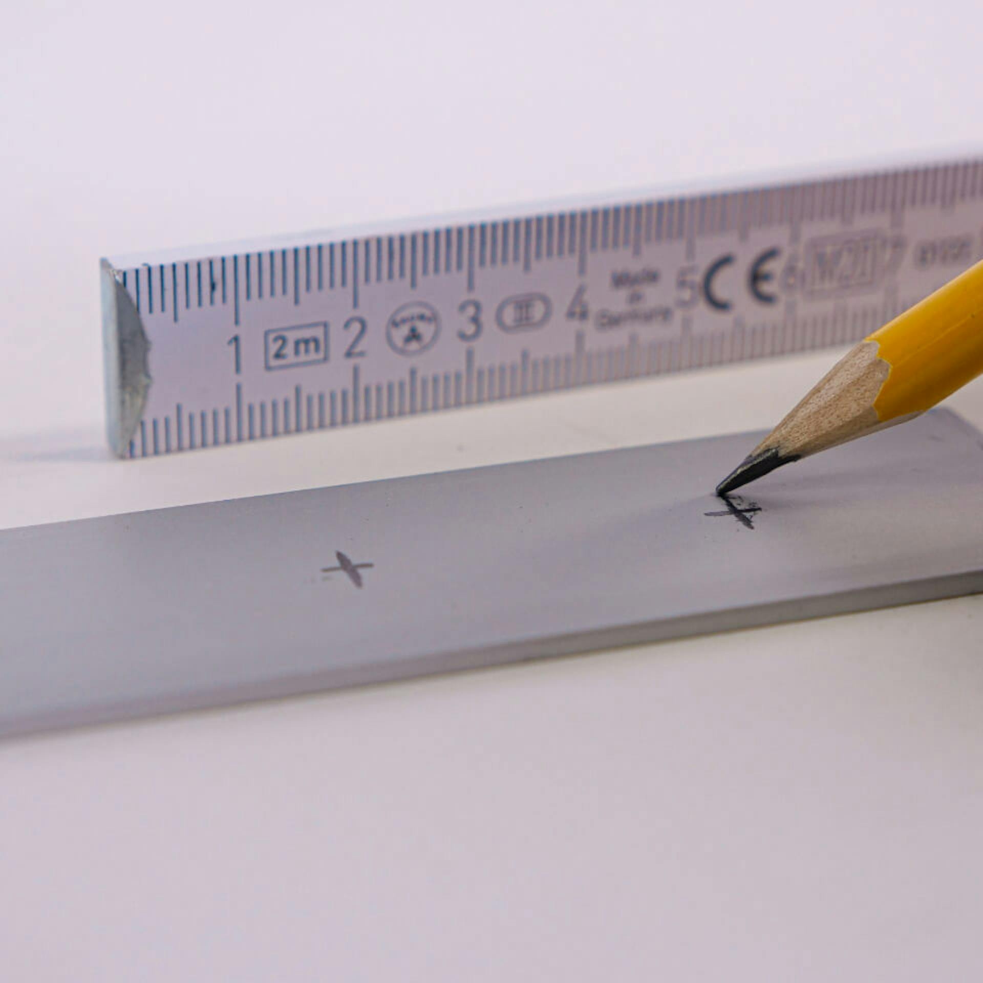 Ceruzával rajzolnak egy jelet egy alumíniumprofilra. Mellette egy mérőléc.