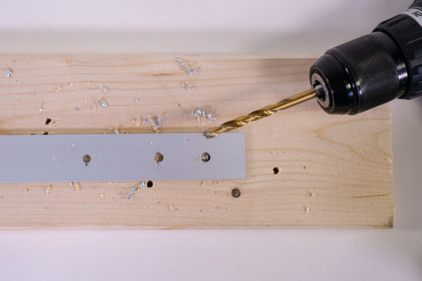 Egy személy akkus fúróval lyukakat fúr egy alumíniumprofilra. Egy fadeszka szolgál alapként.