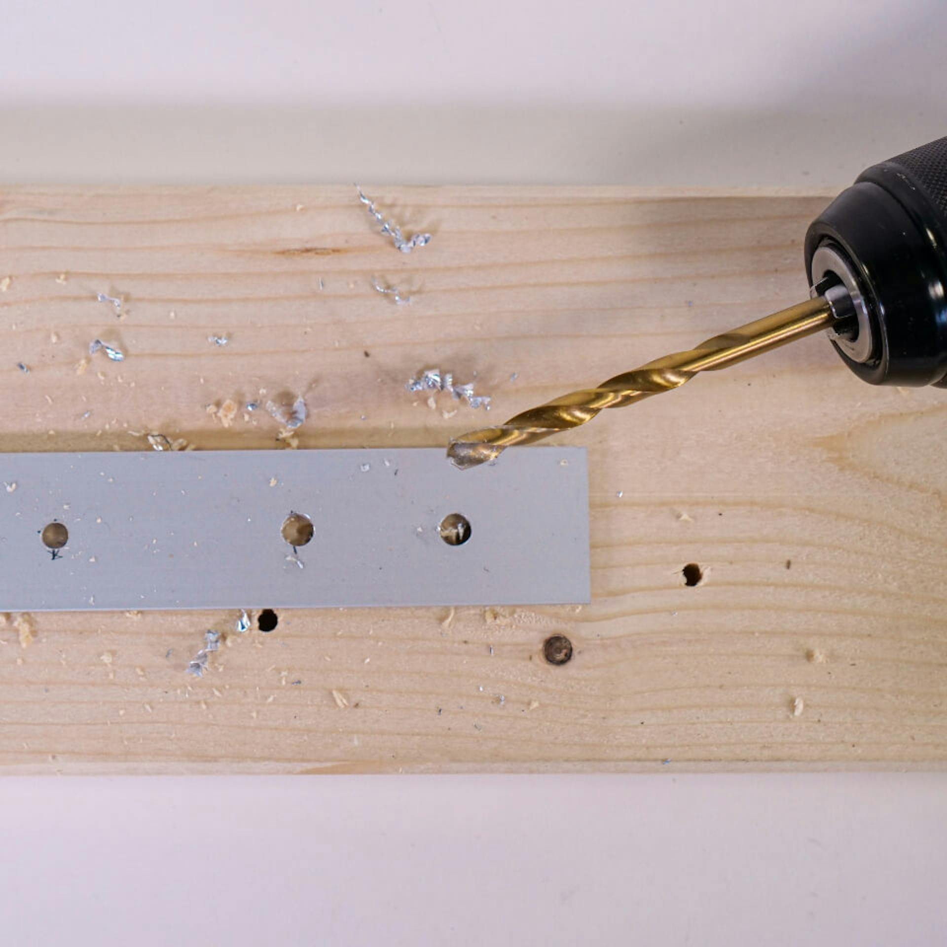 Egy személy akkus fúróval lyukakat fúr egy alumíniumprofilra. Egy fadeszka szolgál alapként.