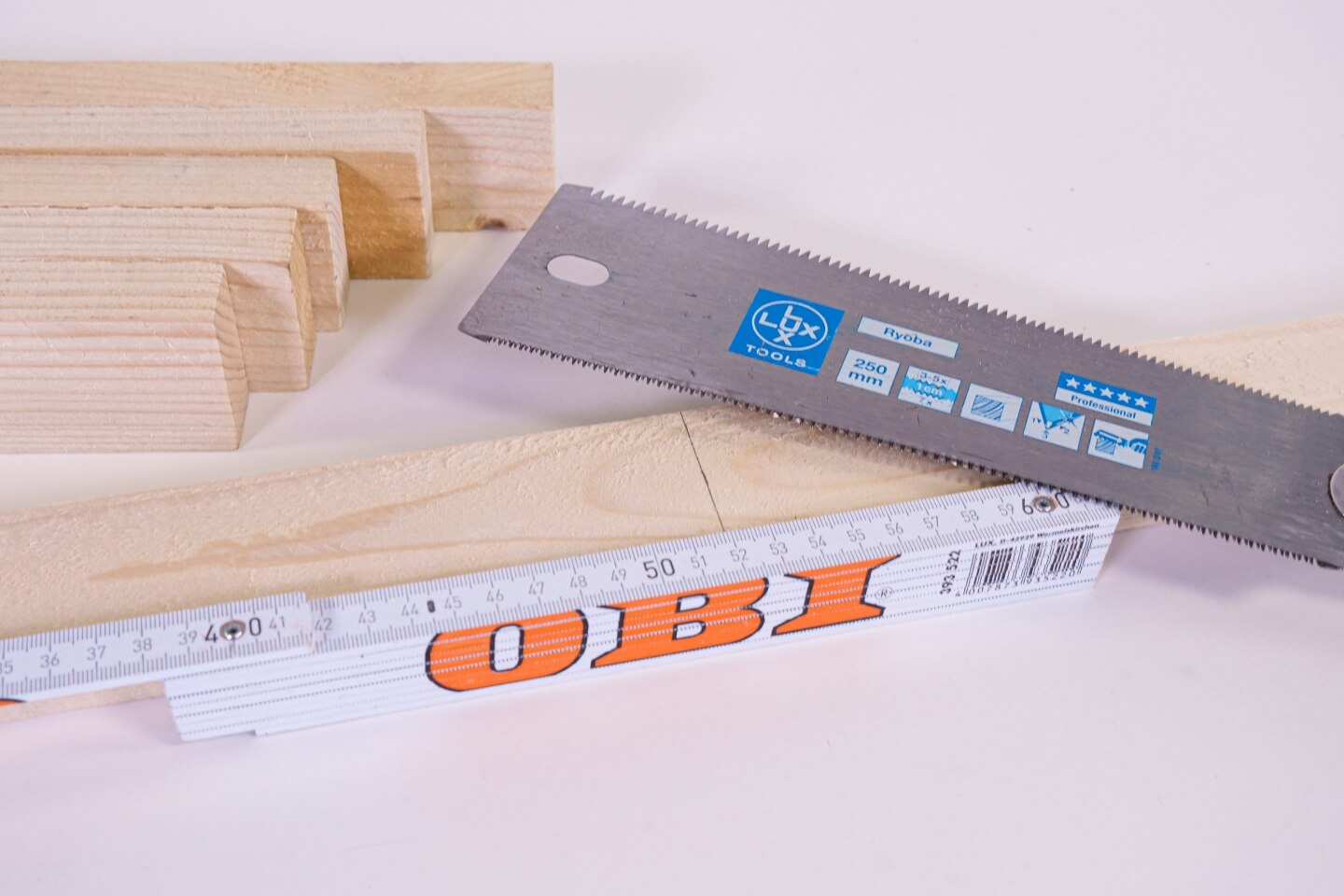 Egy asztalon egy jelekkel ellátott léc fekszik. Mellette egy mérőléc és egy kézifűrész.