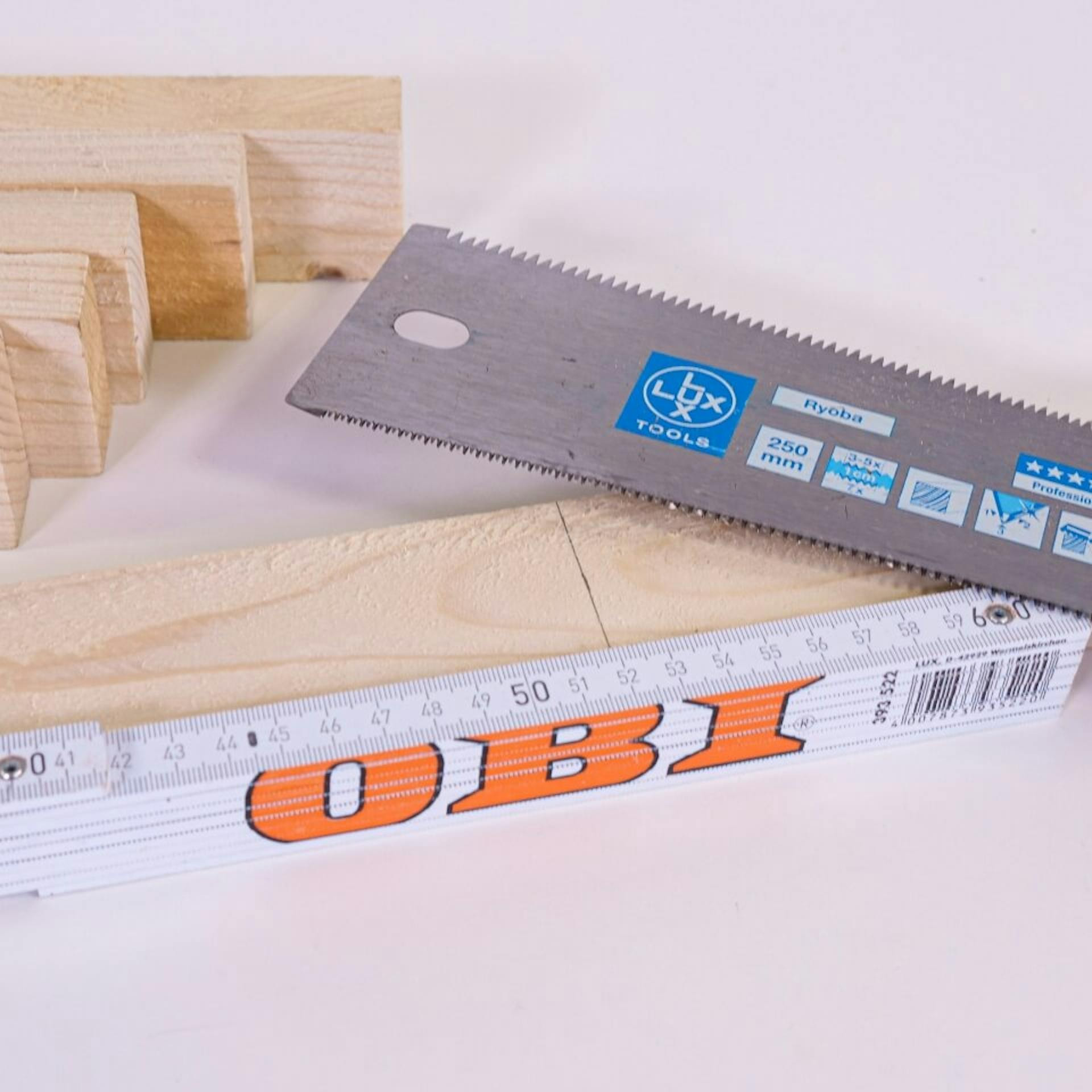 Egy asztalon egy jelekkel ellátott léc fekszik. Mellette egy mérőléc és egy kézifűrész.