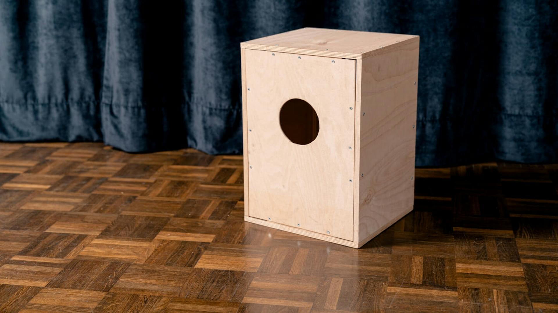 Auf einem Holzboden steht eine Holzkiste, in der Vorderseite ist ein kreisrundes Loch zu sehen. Im Hintergrund ist ein dunkler Samtvorhang zu sehen.