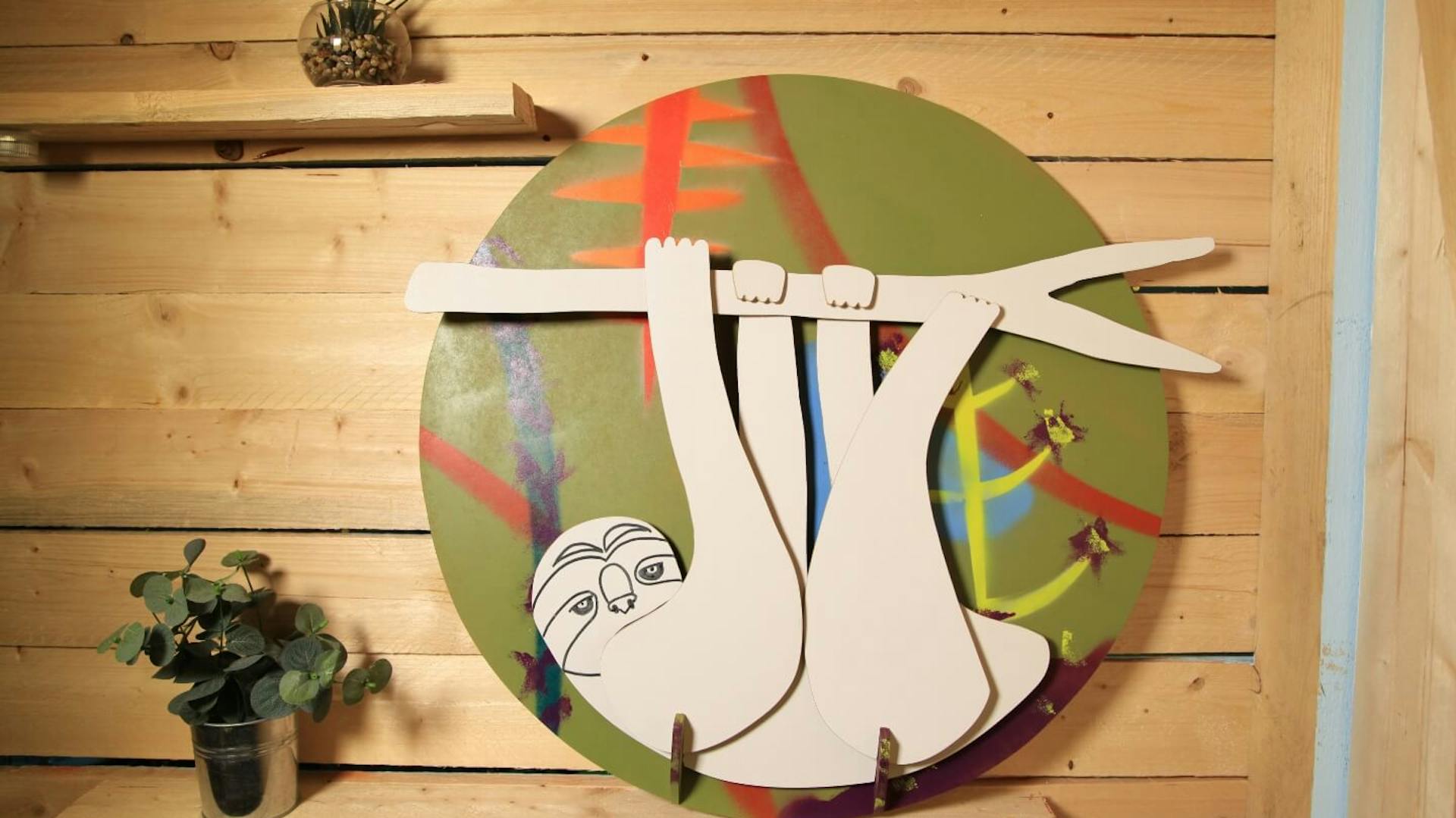 An einer Holzwand ist eine runde bunt bemalte Platte befestigt. Auf der Platte sind Holzelemente in Form eines Faultiers montiert.
