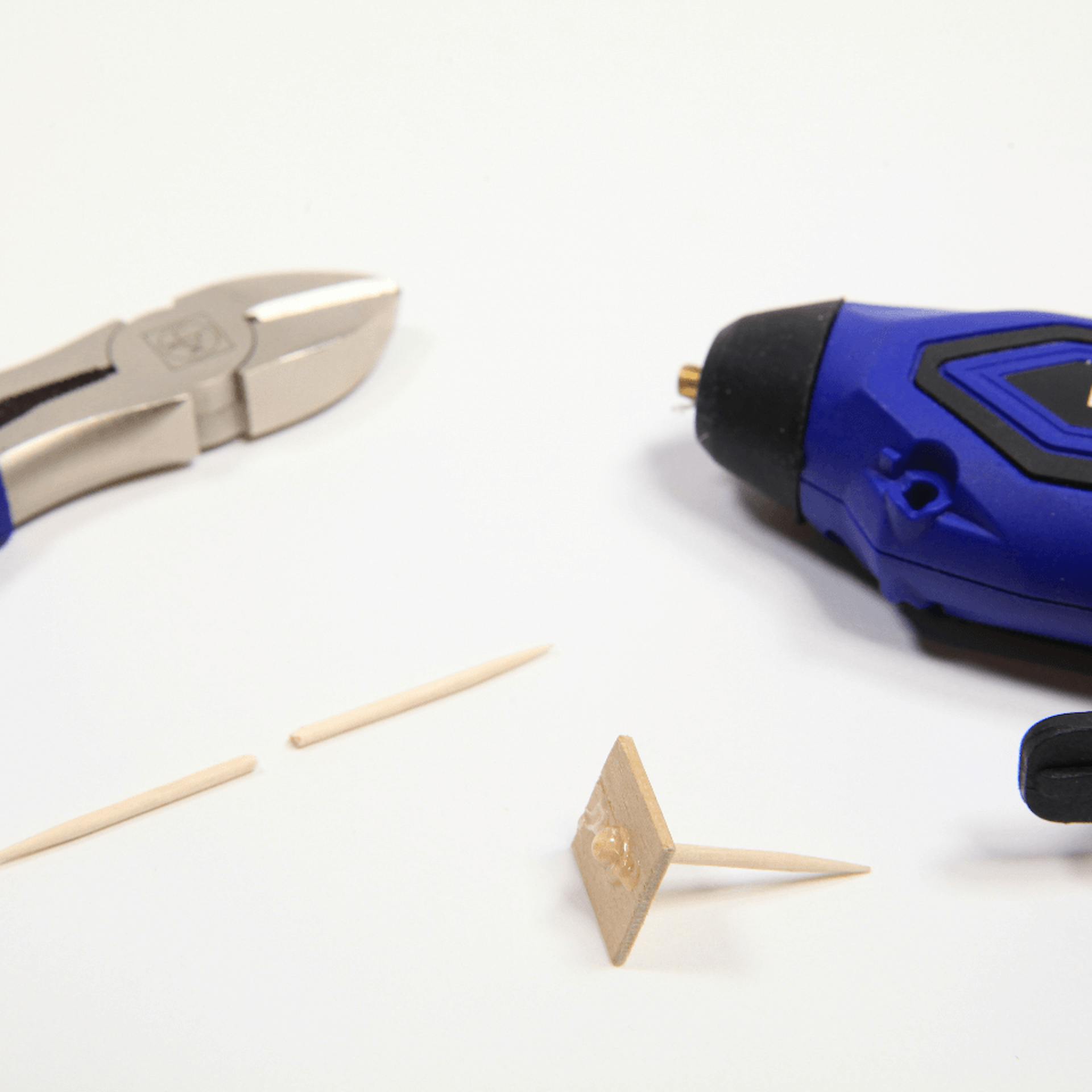 Eine Zange, ein Akkubohrer, ein zerteilter Zahnstocher, ein zerteilter Zahnstocher steckt in einem Holzplättchen.