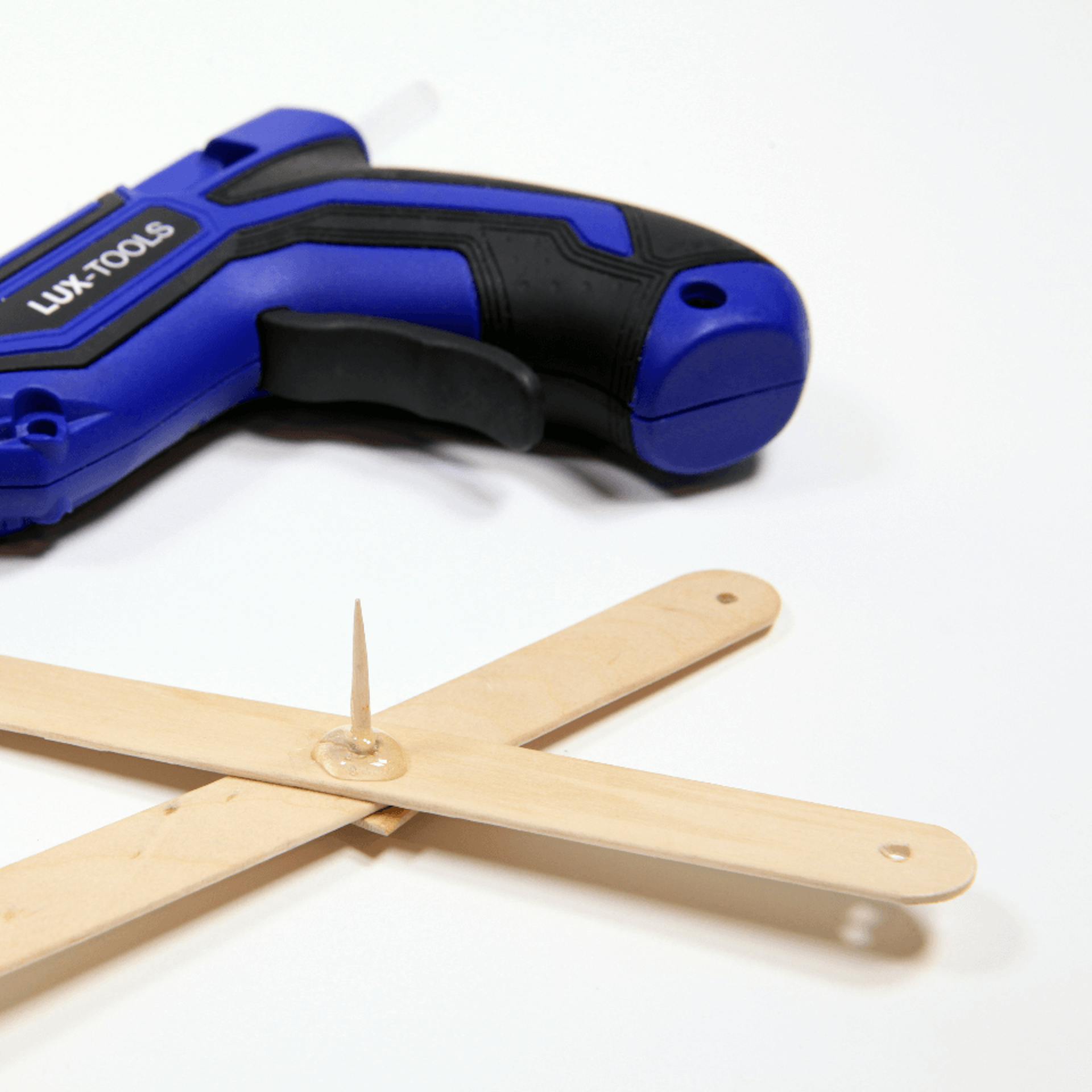 Zwei Holzstücke verbunden mit einem Zahnstocher, dahinter liegt eine Heißklebepistole.
