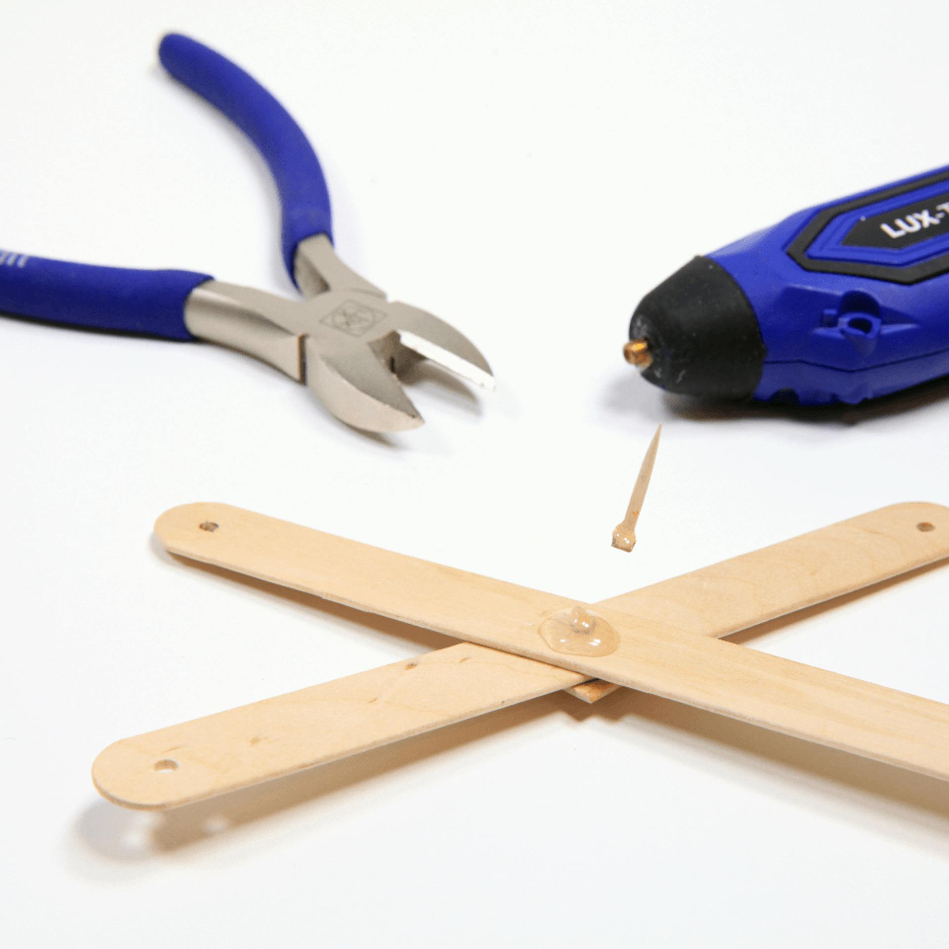 Zwei gekreuzte Holzstücke, eine abgetrennte Zahnstocherspitze, eine Zange und eine Heißklebepistole.