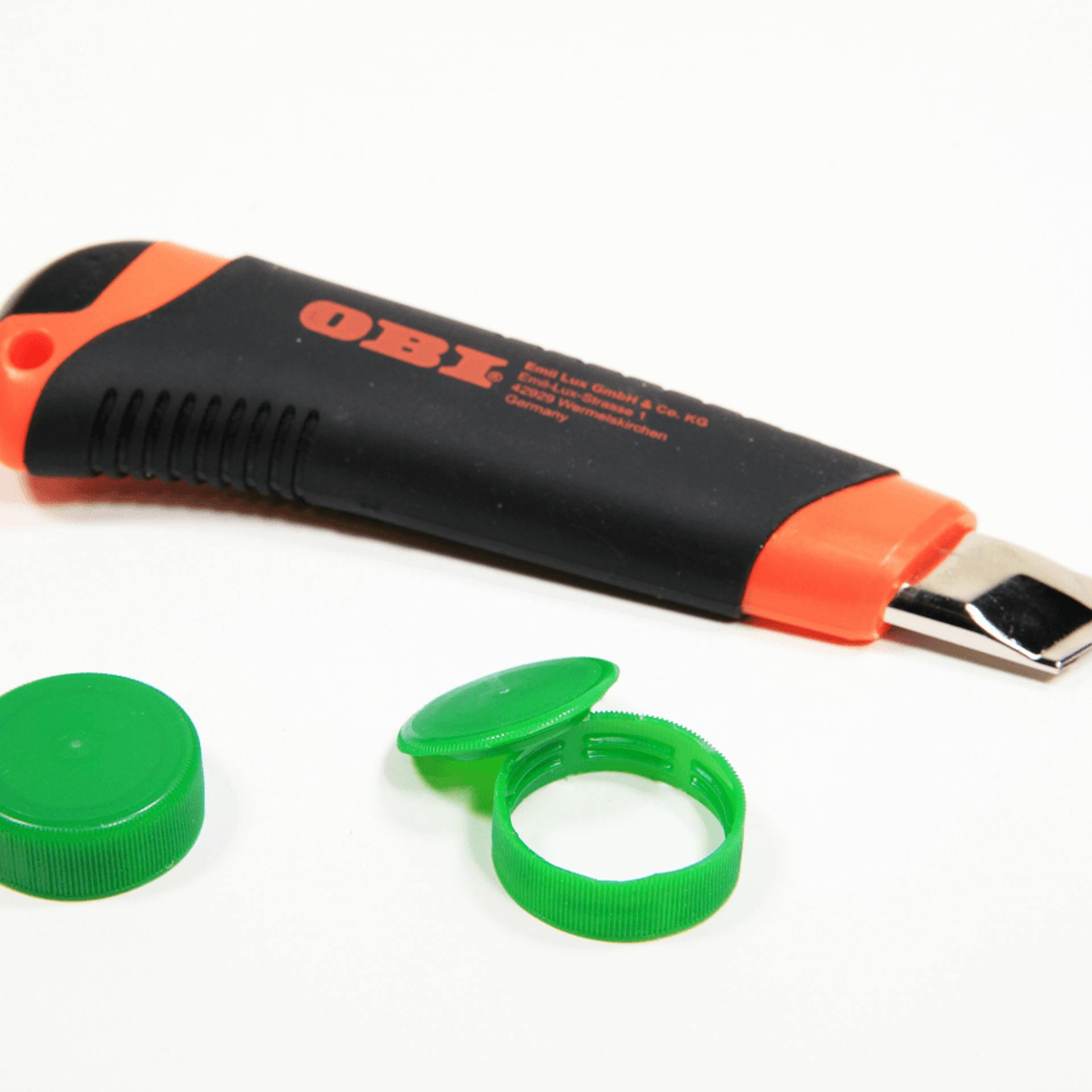 Zwei Flaschendeckel, bei dem einen ist die obere Kappe abgeschnitten, dahinter liegt ein Cuttermesser.