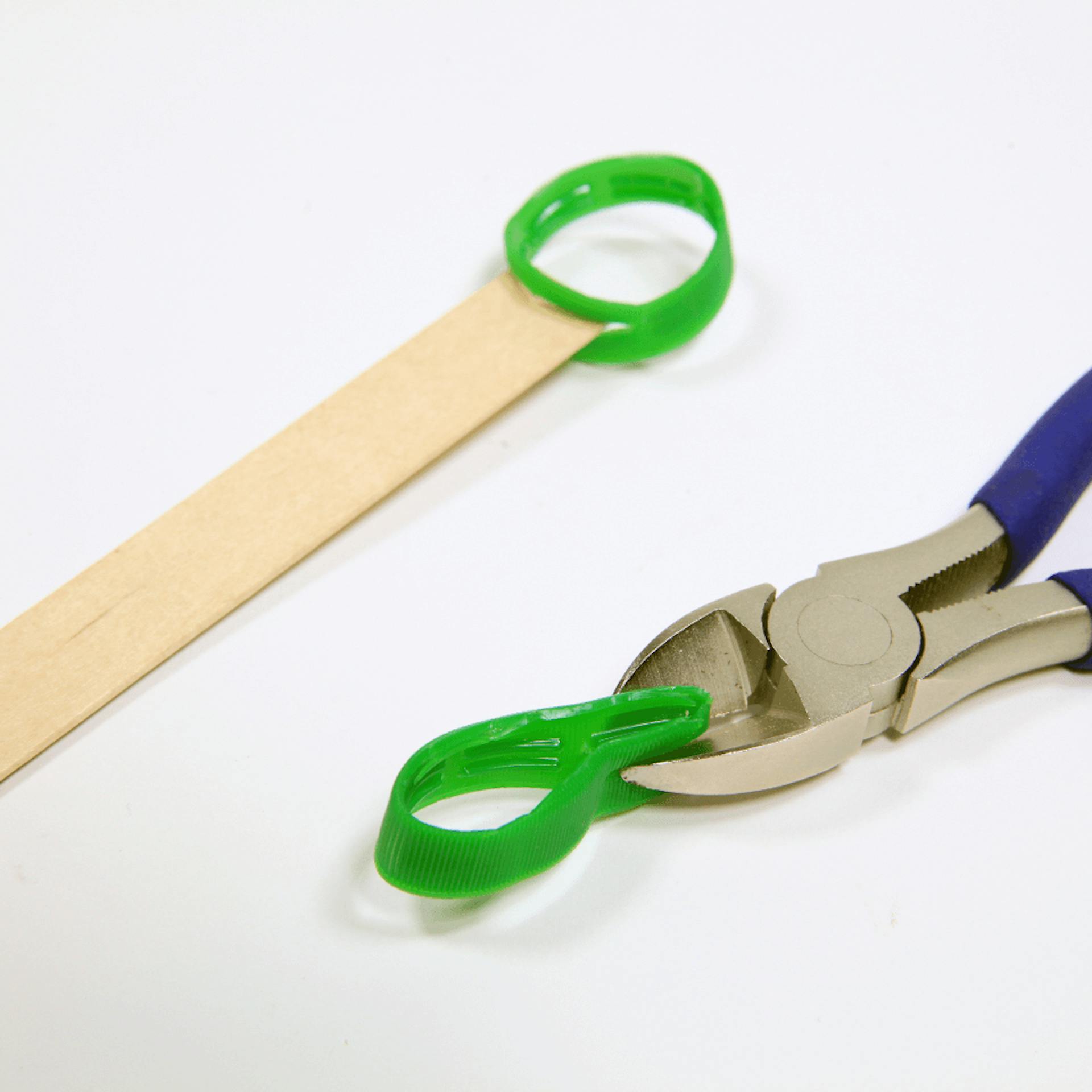Ein Holzspatel mit einem festgeklebten Ring. Eine Zange, die einen Schlitz ein einen zweiten Ring schneidet.