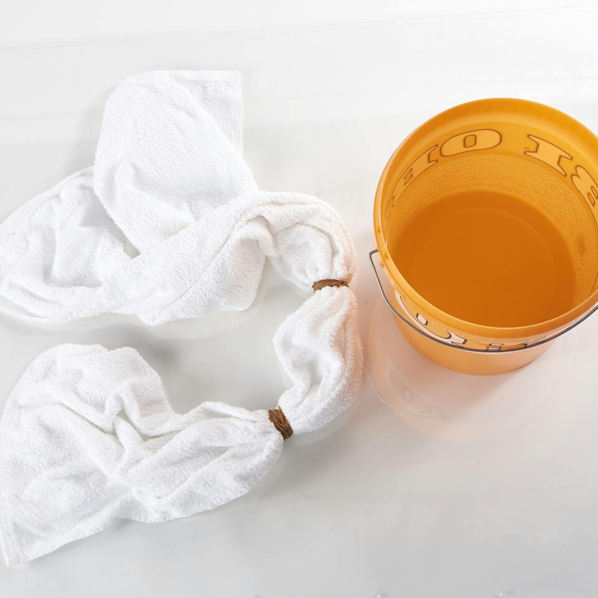 Es ist ein weißes Handtuch zu sehen, welches an zwei Stellen zusammengebunden ist. Daneben steht ein orangefarbener Eimer.