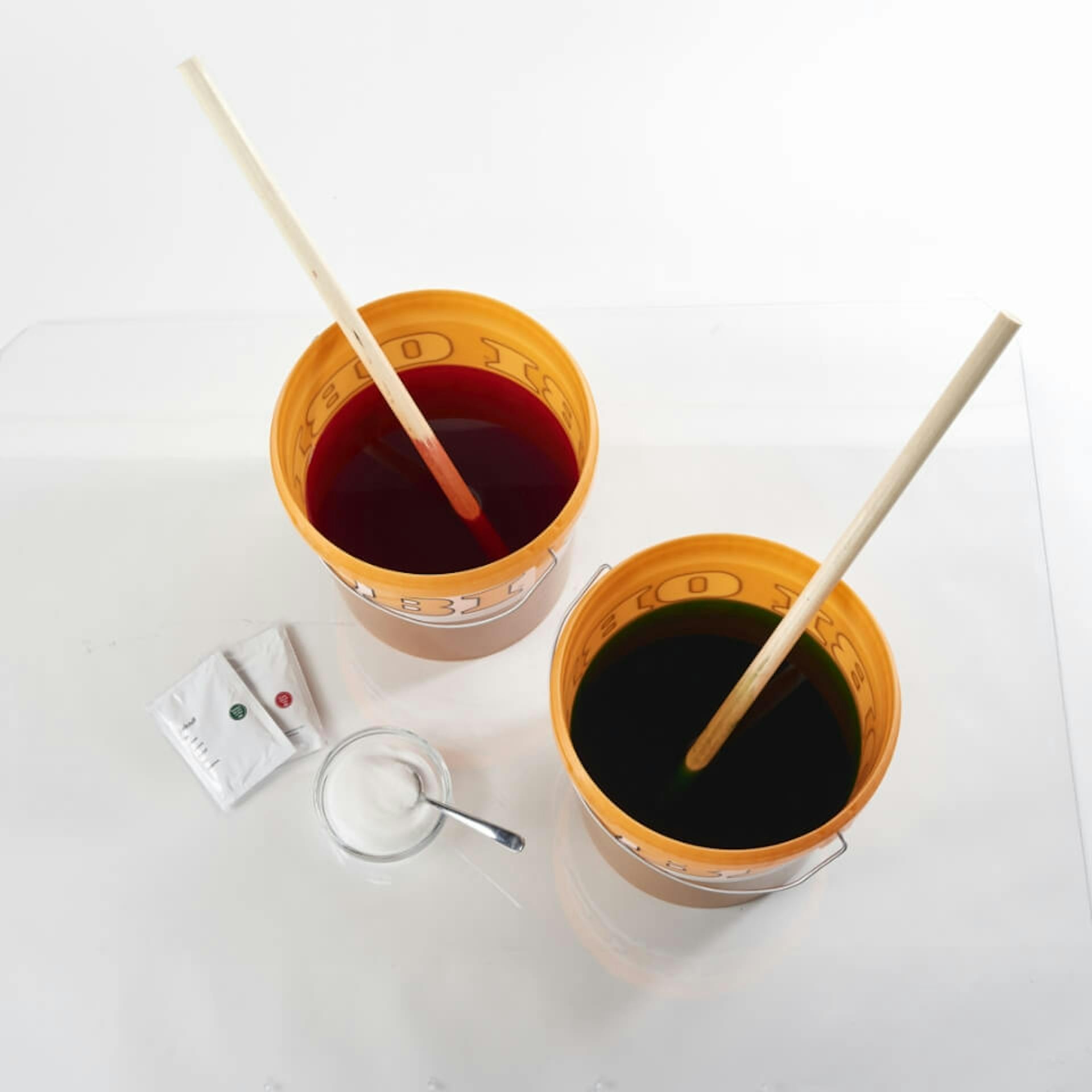 Es sind zwei orangefarbene Eier zu sehen, welche jeweils mit Textilfarbe gefüllt sind. In jedem Eimer steht zudem ein Holzstab. Neben den Eimern steht eine Schale mit Kochsalz. Daneben liegen zwei kleine weiße Tüten.