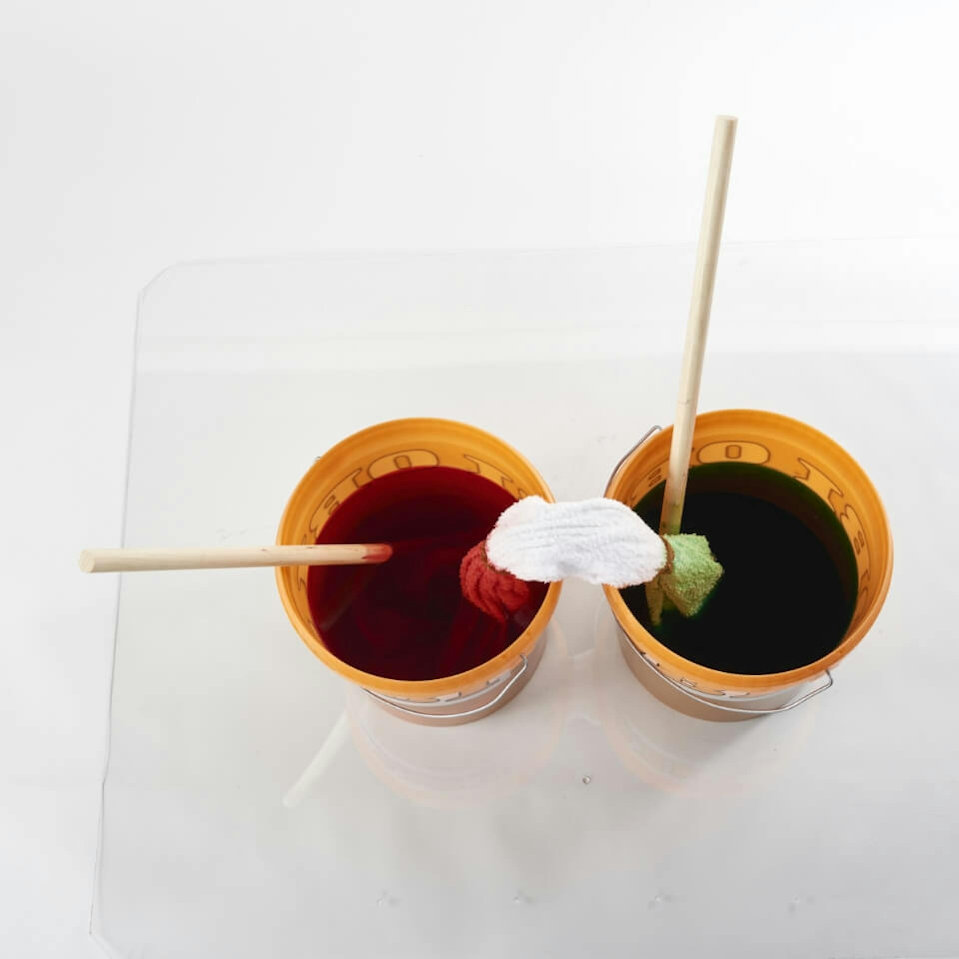 Es sind zwei orangefarbene Eimer zu sehen, welche jeweils mit Textilfarbe befüllt sind. In jedem Eimer steht zudem ein Holzstab. Es ist ein weißes Handtuch zu sehen, welches mit je einem Ende in einem Eimer hängt.