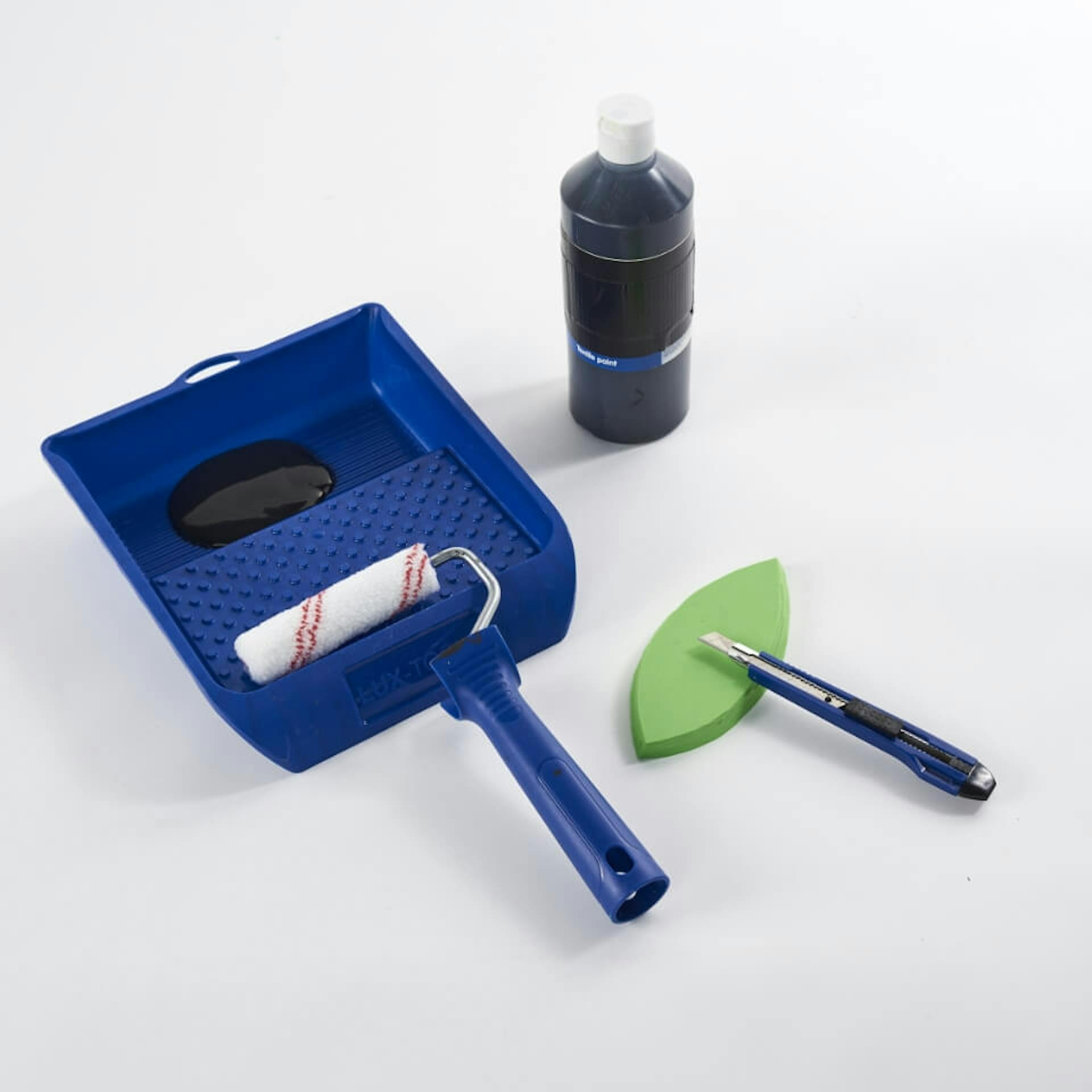 Es ist ein Farbroller in einer blauen Farbwanne zu sehen. Daneben steht schwarze Textilfarbe. Davor liegen grüne Moosgummiplatten in Tropfenform. Auf dem Moosgummi liegt ein Cutter.