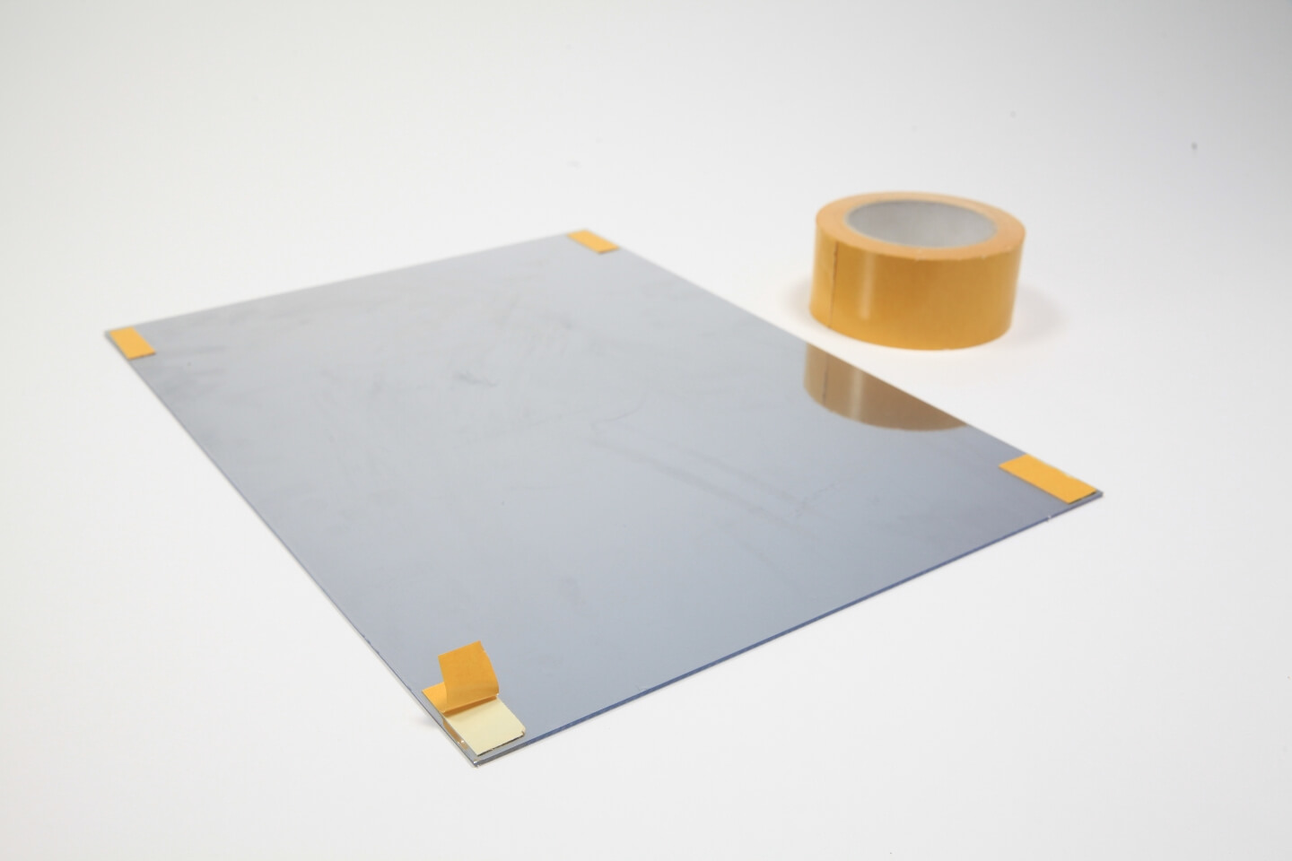 Plexiglas mit Spiegelfolie liegt auf einem weißen Untergrund. In jeder Ecke der Scheibe sind kurze Streifen von Teppichklebeband befestigt.