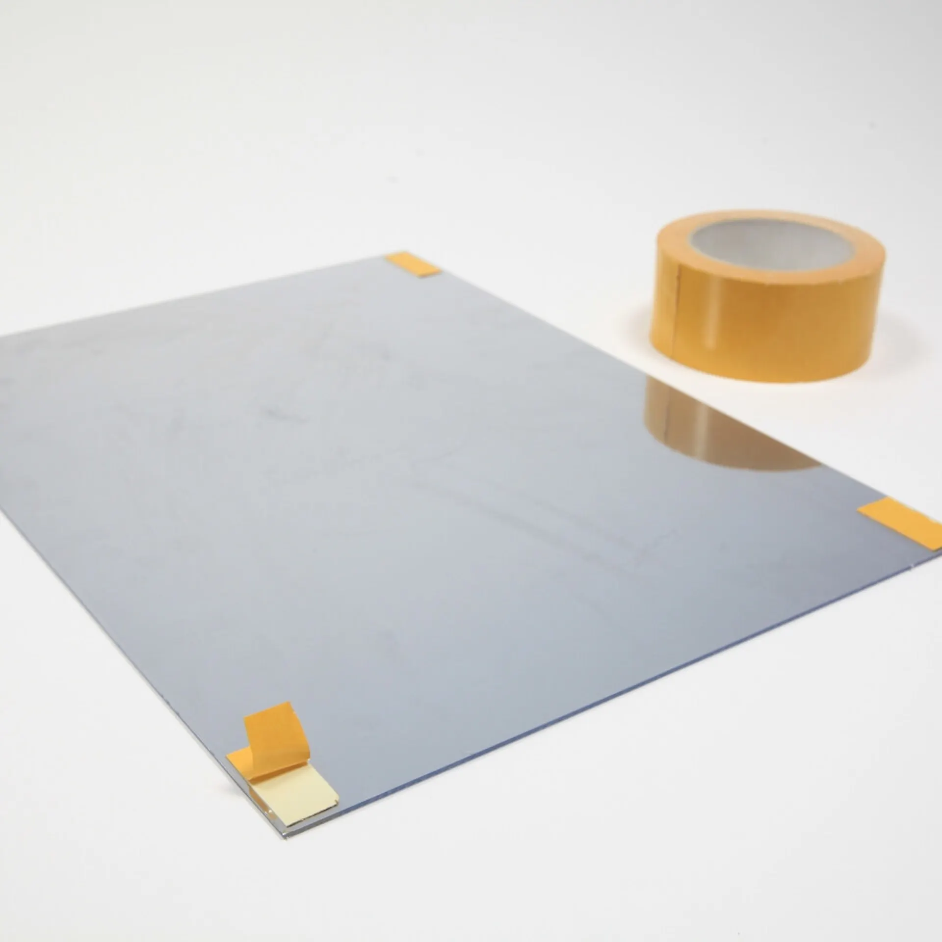 Plexiglas mit Spiegelfolie liegt auf einem weißen Untergrund. In jeder Ecke der Scheibe sind kurze Streifen von Teppichklebeband befestigt.