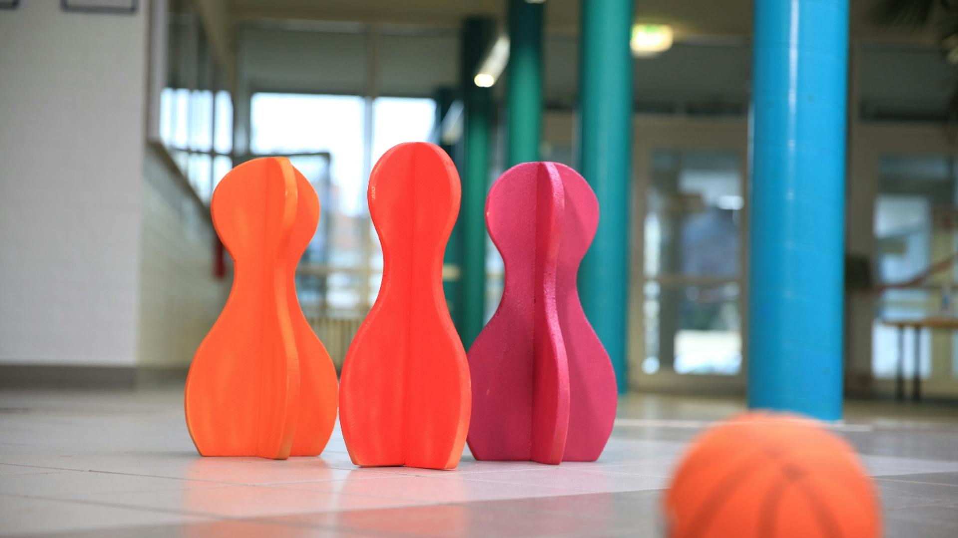 Es sind drei Kegel in verschiedenen Rot-Tönen zu sehen. Davor liegt ein kleiner Ball in Basketball-Optik. Im Hintergrund sind mehrere blaue Säulen zu sehen.