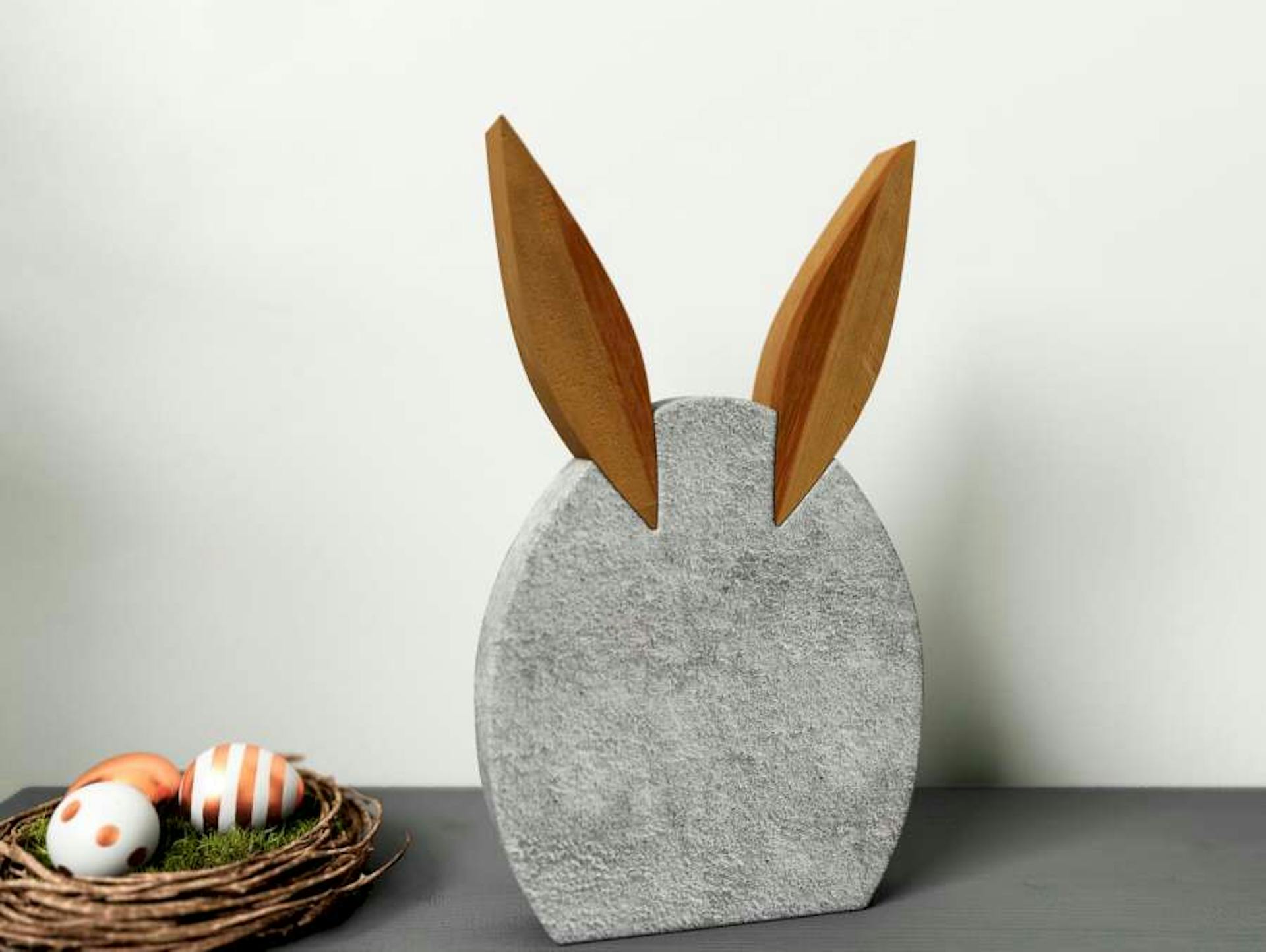 Osterdeko in Form von Hase aus Beton steht auf Kommode neben kleinem Osternest mit bunten Eiern.