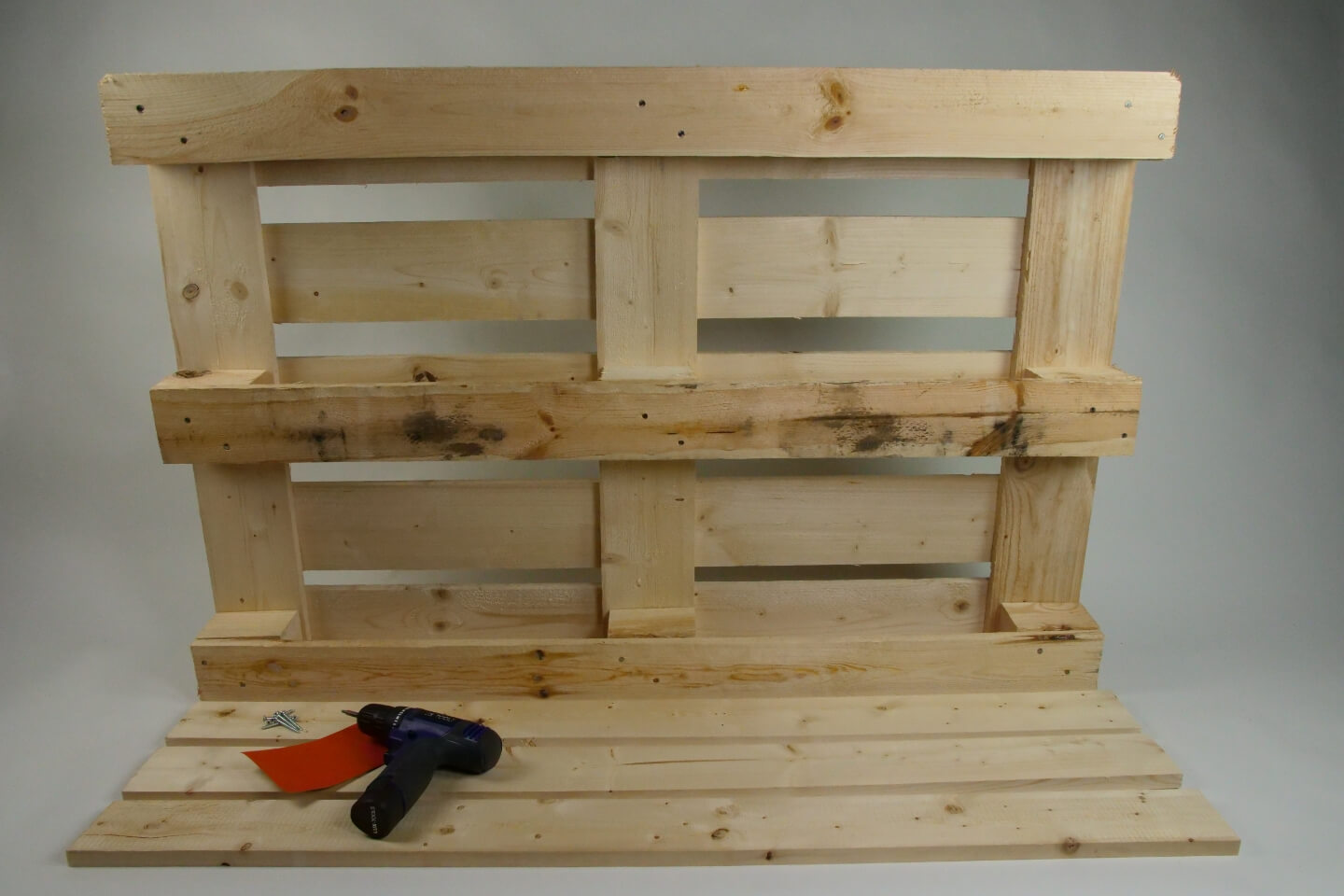 Es ist die Rückseite einer Holzpalette zu sehen. Davor liegen drei identische Holzbretter. Auf diesen liegt Schleifpapier sowie ein Akkubohrer.