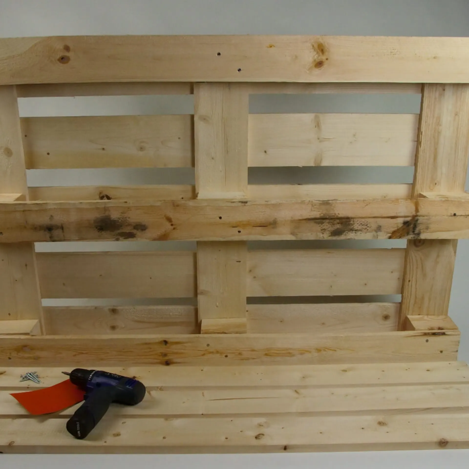 Es ist die Rückseite einer Holzpalette zu sehen. Davor liegen drei identische Holzbretter. Auf diesen liegt Schleifpapier sowie ein Akkubohrer.