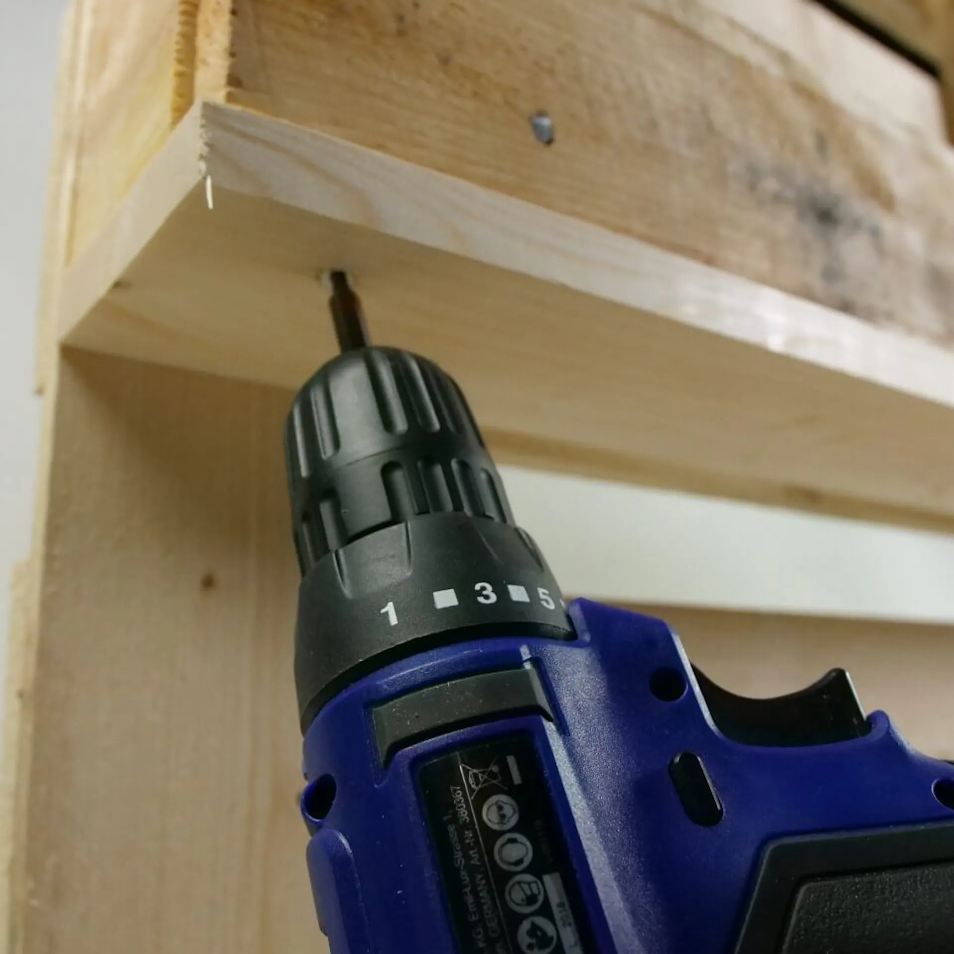 Es ist eine Nahaufnahme einer Holzpalette zu sehen. Mit einem Akkubohrer wird ein Brett an dieser befestigt.