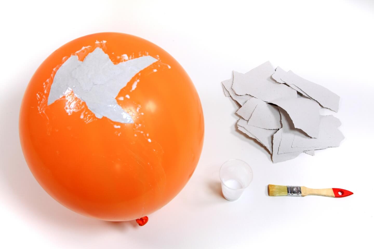Zu sehen ist ein aufgepusteter Luftballon, auf dem bereits einige Papierschnipsel mit Leim aufgeklebt sind. Daneben liegen zerrissene Papierstücke, eine Leimmischung in einem Becher und ein Flachpinsel.