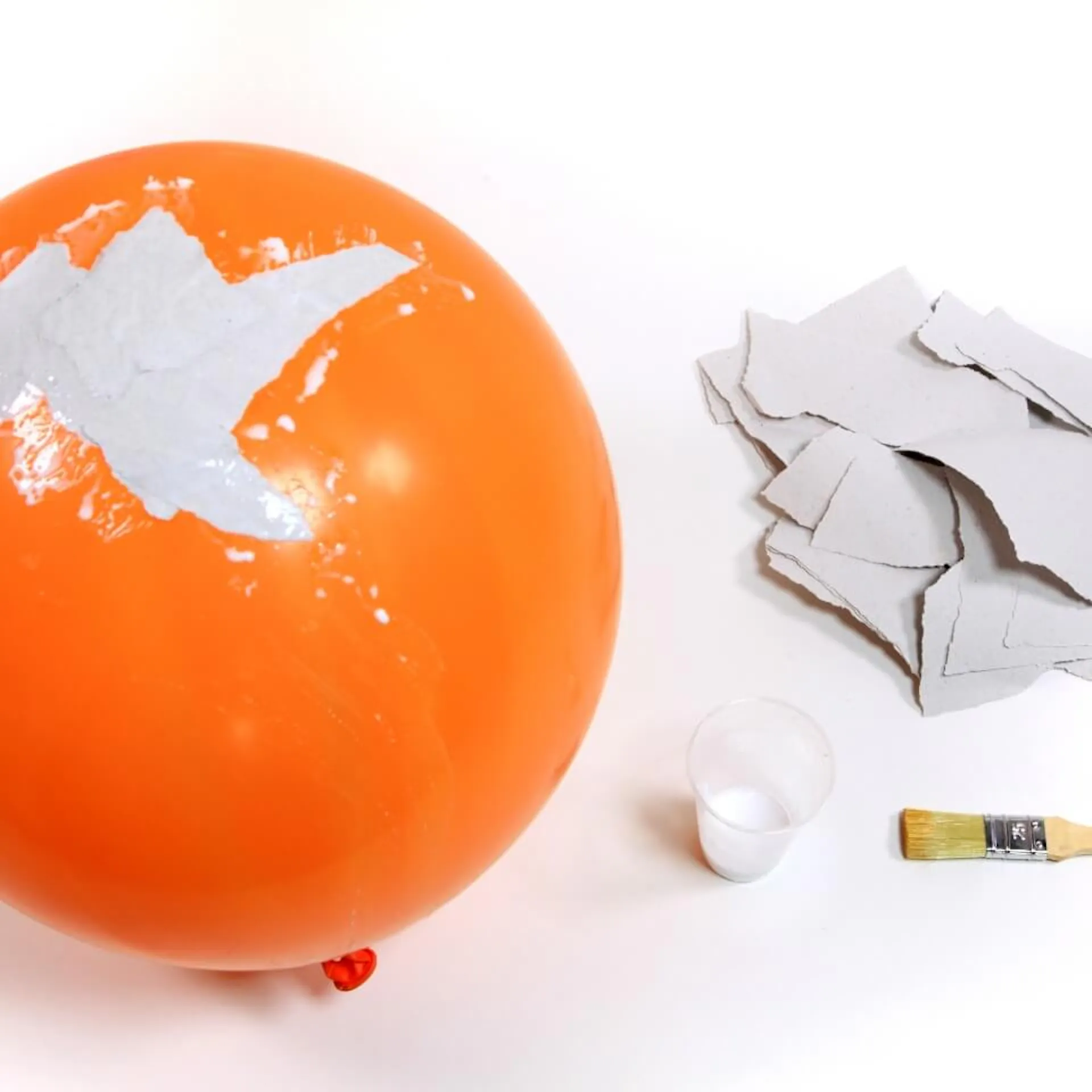 Zu sehen ist ein aufgepusteter Luftballon, auf dem bereits einige Papierschnipsel mit Leim aufgeklebt sind. Daneben liegen zerrissene Papierstücke, eine Leimmischung in einem Becher und ein Flachpinsel.